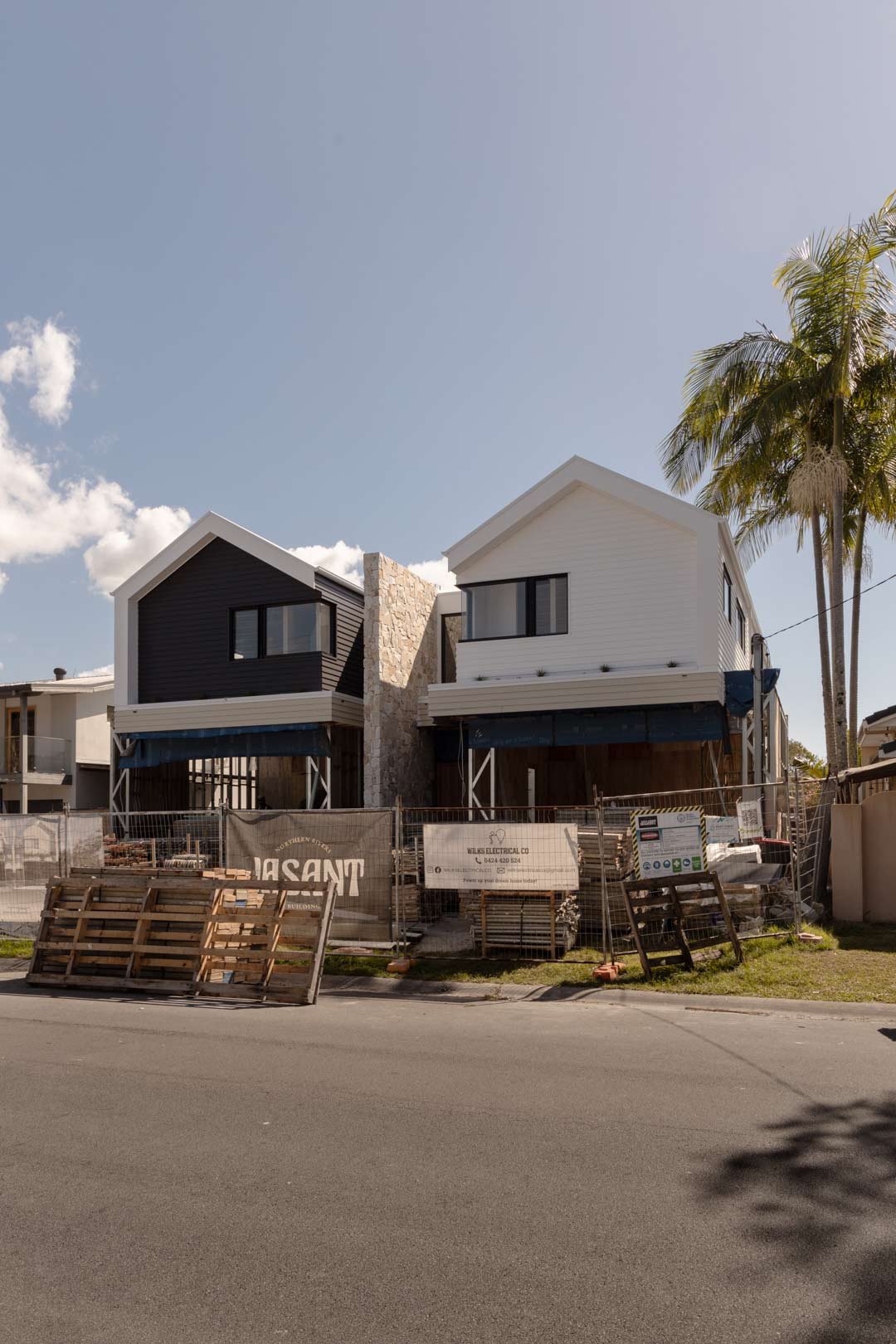 Two modern houses under construction with scaffolding and construction materials in front, along a paved street, with palm trees and a blue sky in the background.