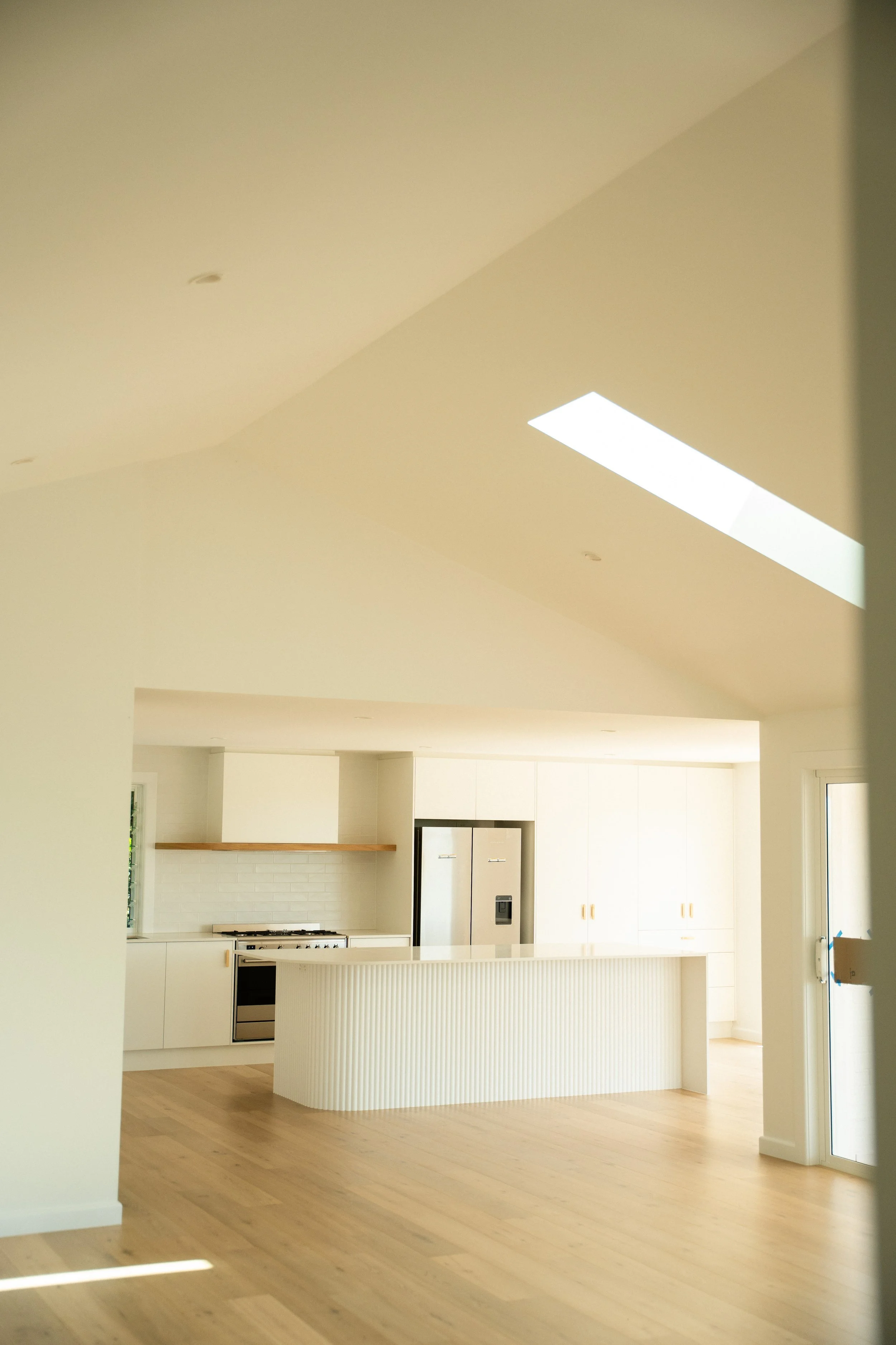 A spacious, modern kitchen with white cabinets, a stainless steel refrigerator, a built-in oven, a white tiled backsplash, and a kitchen island with a curved, ribbed design. Hardwood floors and a skylight overhead provide natural lighting.