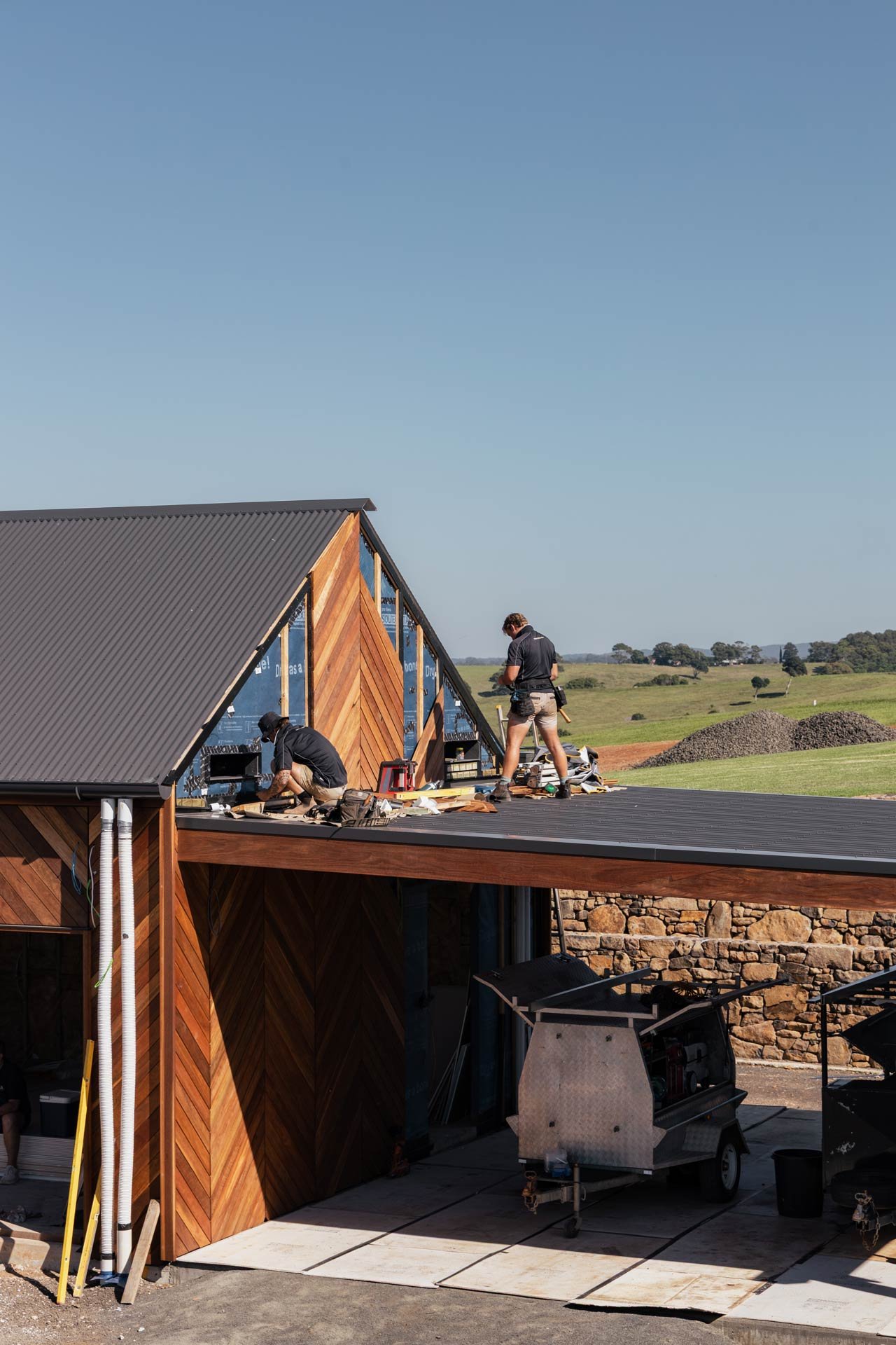 Two workers installing or repairing a wooden building with a steep metal roof, outdoors in a rural area with green fields and trees under a clear blue sky.