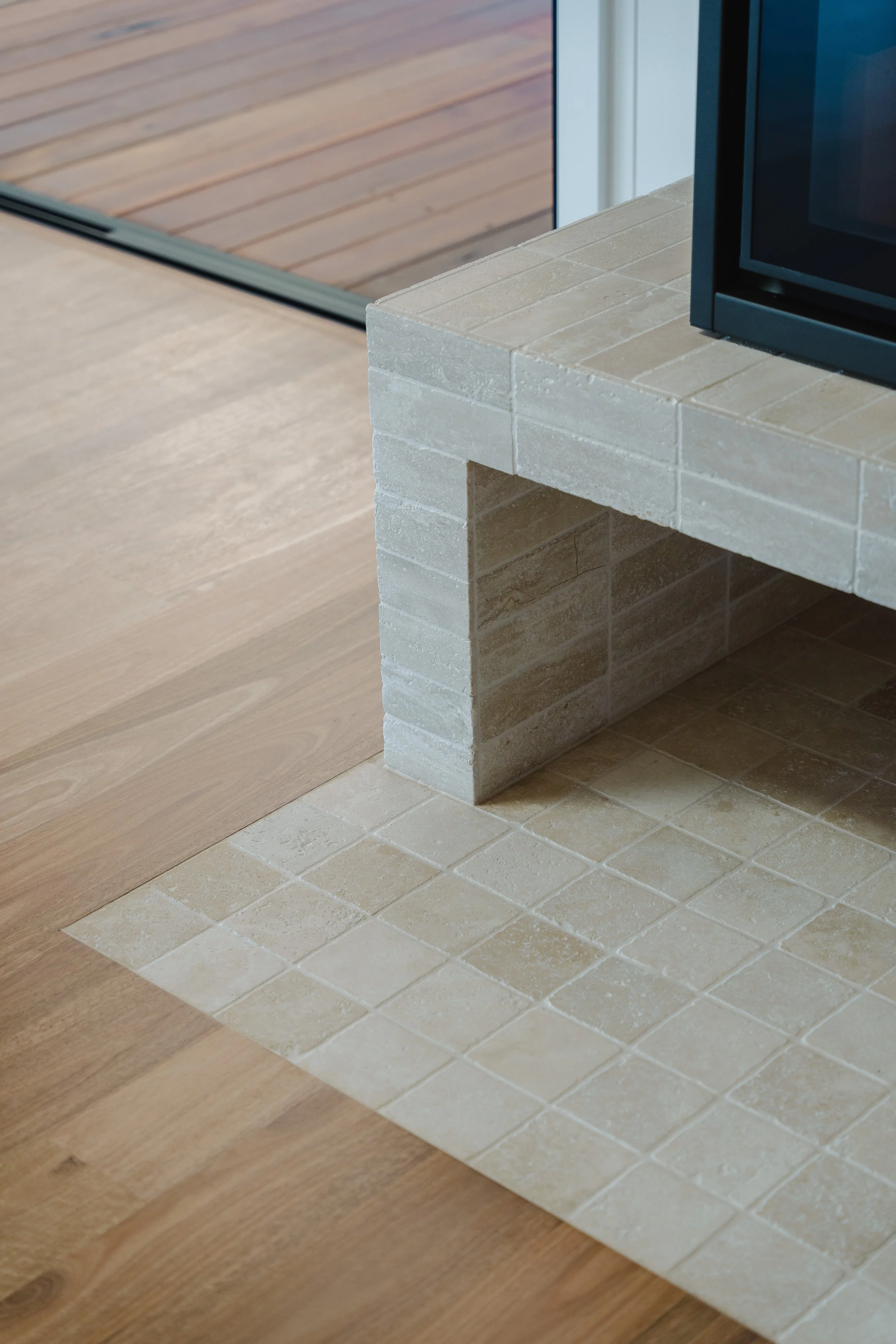 Close-up view of a fireplace with a black frame and a beige brick hearth. The hearth extends onto a light wood floor, with beige tiles arranged in a rectangular pattern in front of it. The background shows a sliding glass door with a wooden deck outside.