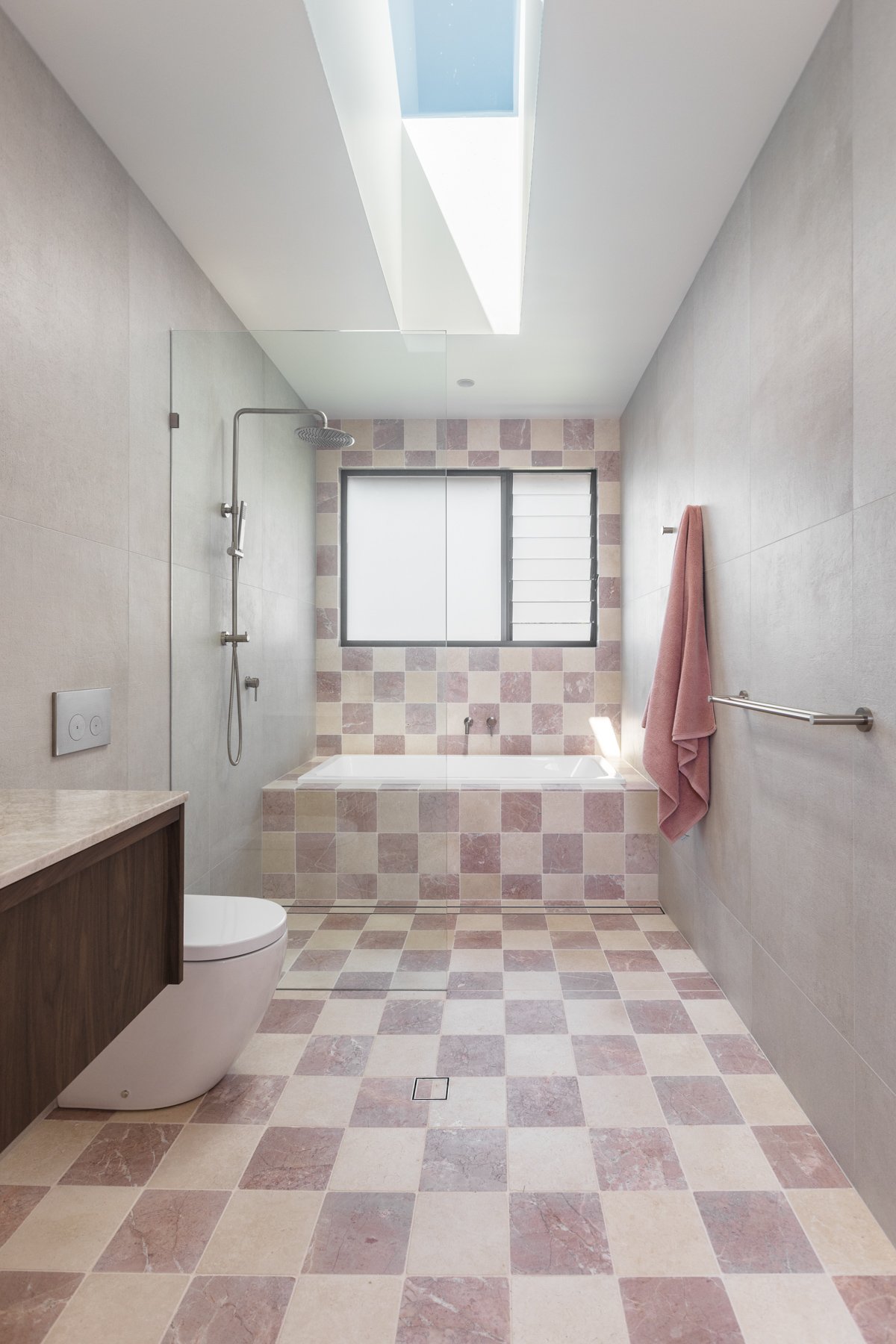Modern bathroom with a bathtub, a walk-in shower with a ceiling-mounted showerhead, a window with frosted glass and louvers, a toilet, a pink towel hanging on a towel rack, beige and pink checkered floor and wall tiles, a skylight, and a wooden vanity