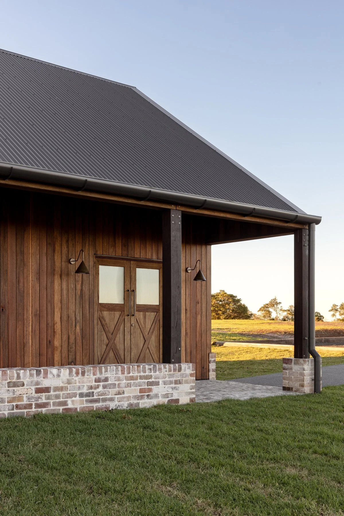 A wooden barn with a metal roof and brick foundation, set in a grassy landscape with trees in the distance during sunset.