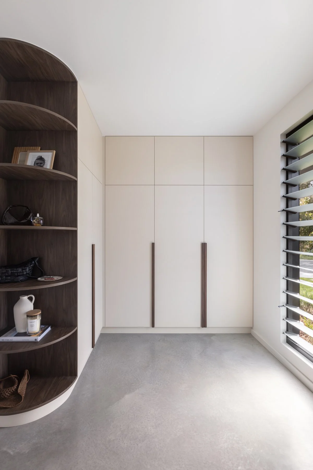 Minimalist interior with built-in white wardrobe, rounded wooden shelving unit on the left with decorative items, and a large window with adjustable louvered blinds on the right, letting in natural light.