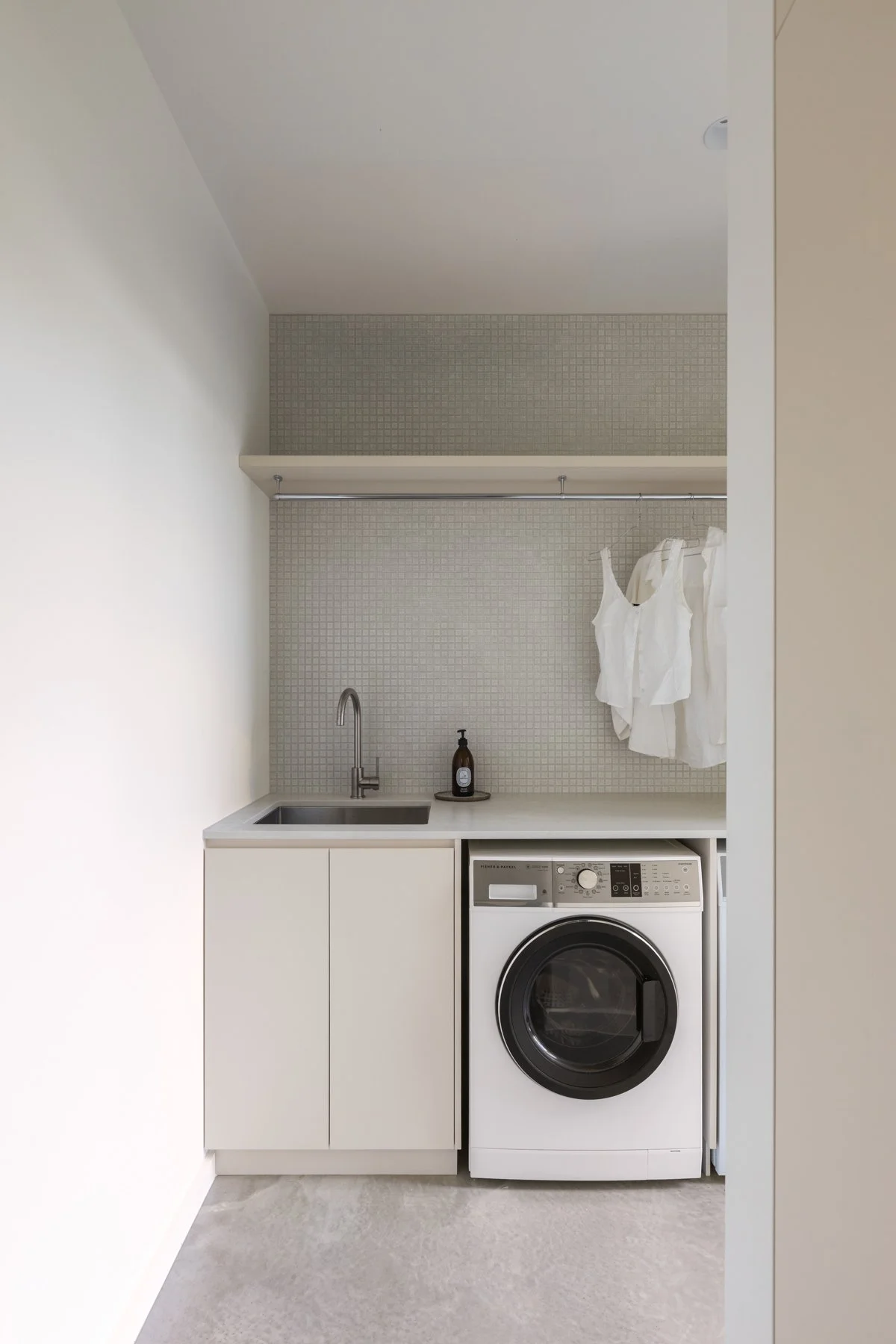 A small laundry area with a white washer and a cabinets beneath a white countertop. Above the counter, there is a small sink with a silver faucet, a soap dispenser, and a shelf with hanging white dresses. The wall behind features small square tiles, and the space is enclosed by white walls and a white ceiling.