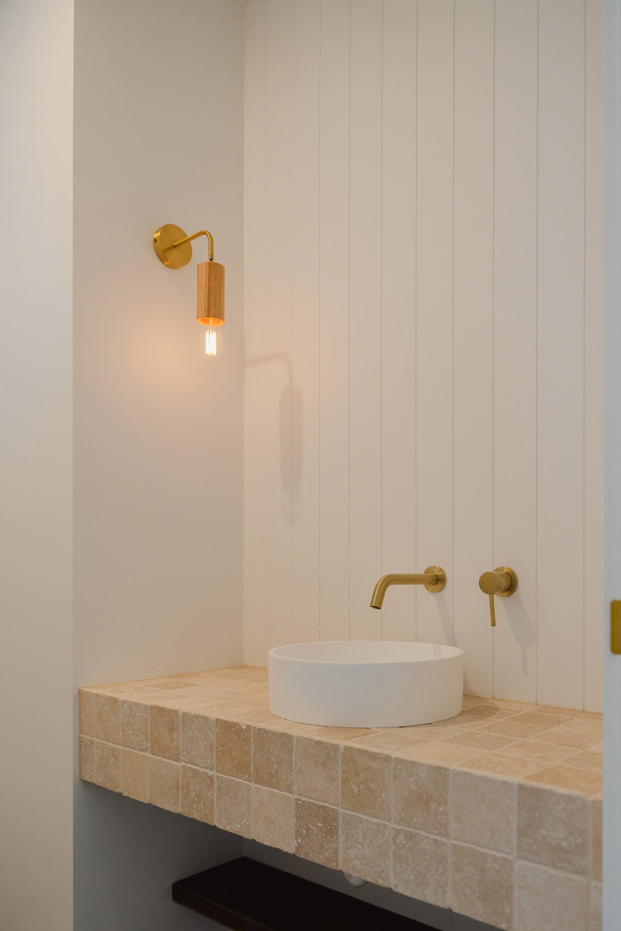 Modern bathroom with a white round vessel sink, gold fixtures, and beige tile countertop and walls.