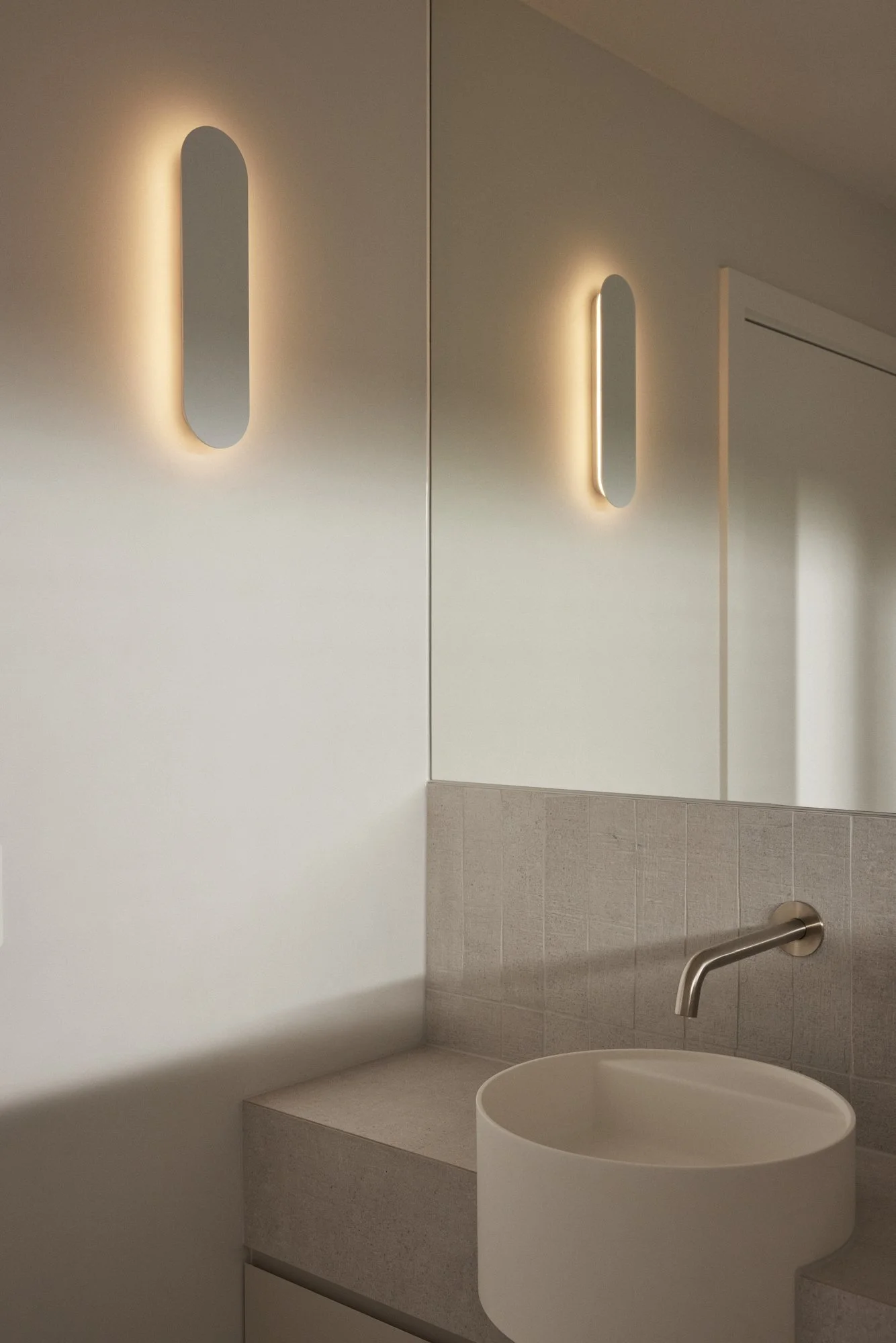 Modern bathroom with oval mirror backlit by soft lighting, beige tiled wall, stainless steel faucet, and a round white vessel sink.
