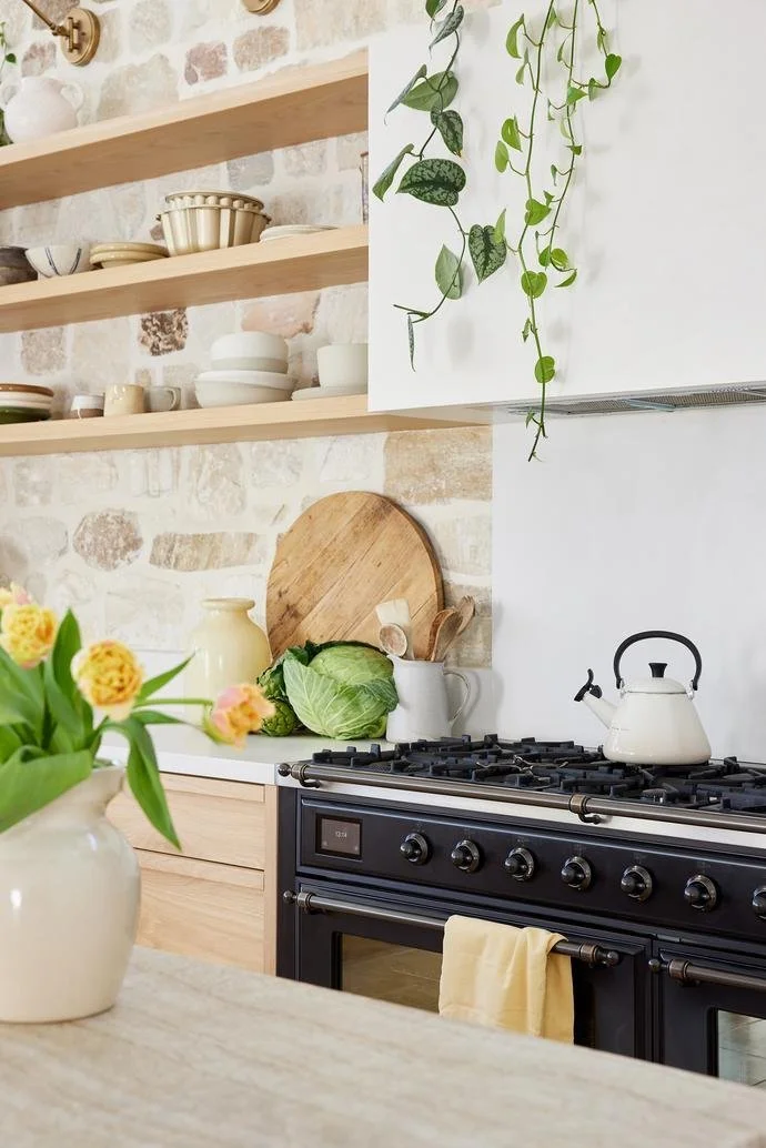 Kitchen with open floating wood shelves, white brick wall, hanging green vines, black stove with a yellow towel, a white teapot, cabbage, a yellow vase, and a round wooden cutting board on the counter, with a vase of yellow flowers in the foreground.