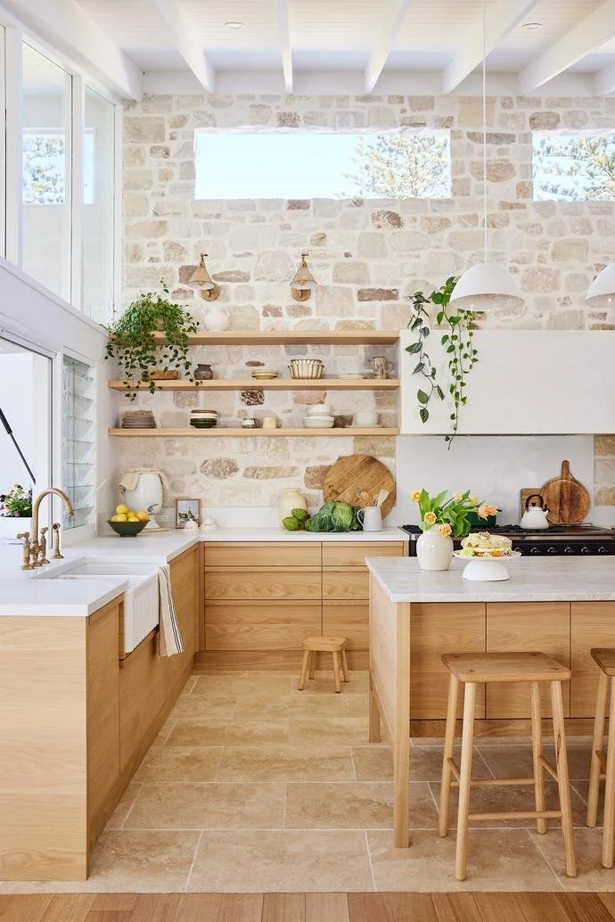 Bright kitchen with white countertops, wooden cabinets, open shelving with dishes, stone wall, and large windows allowing natural light, with stools at an island and decor including plants and kitchenware.