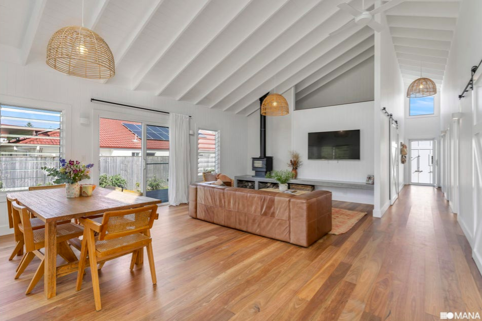 Bright open living and dining area with white vaulted ceiling, wooden flooring, a leather sofa, a mounted flat-screen TV, a wood stove, rattan pendant lights, sliding glass doors with white curtains, and a hallway leading to the front entrance