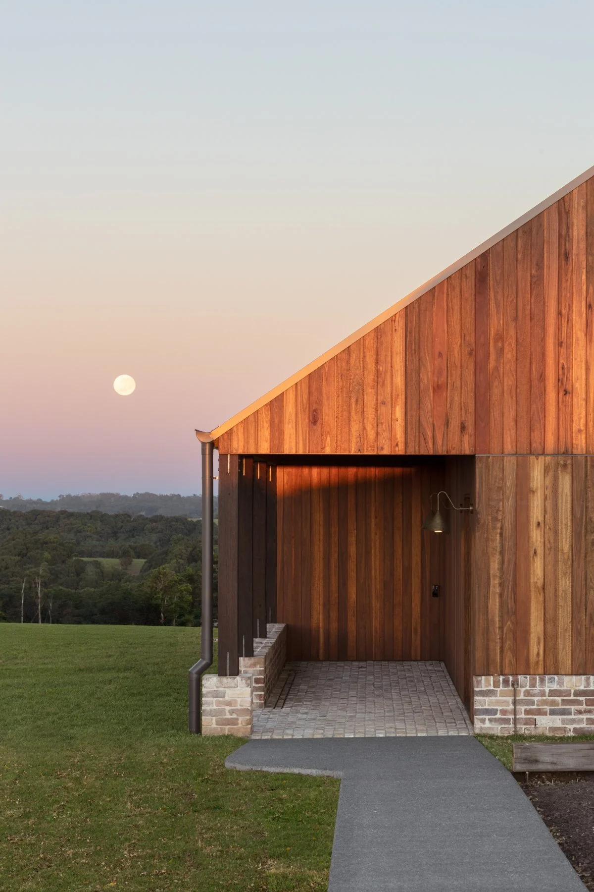 A modern wooden building with a slanted roof, a small porch area with a paved walkway, and a landscape with trees and hills in the background during dusk, with a visible full moon in the sky.