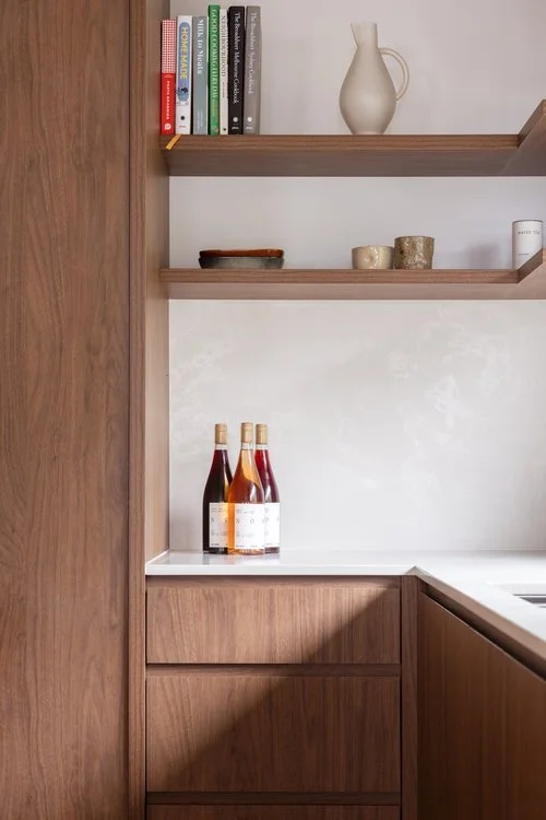 Wooden kitchen shelves with books and decorative items, bottles on a white countertop, and a wooden cabinet below.