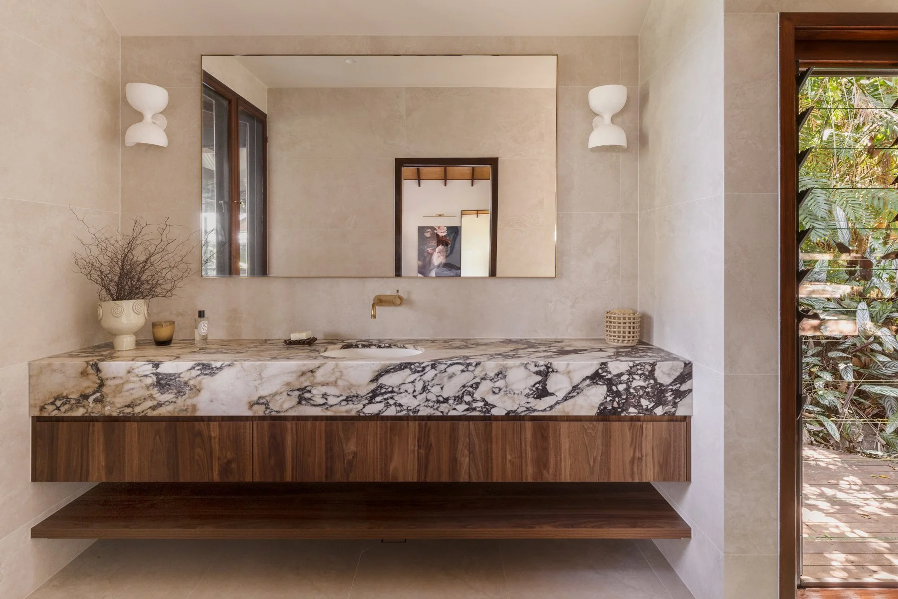 Modern bathroom vanity with a marble countertop, wooden cabinet, large mirror, wall sconces, and a view of lush greenery outside through an open wooden door.