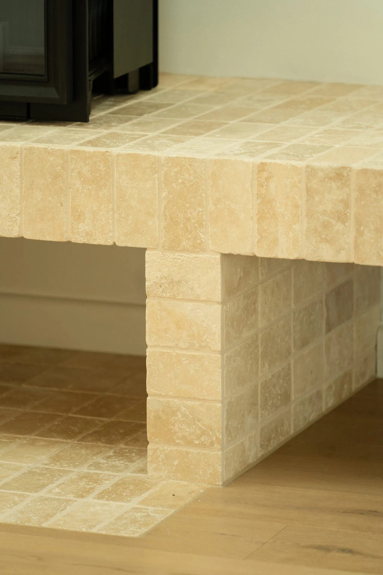 A corner of a beige tiled kitchen counter with a black oven against the wall. The counter has a raised edge, and the floor and lower section of the counter are also tiled in matching beige stones.