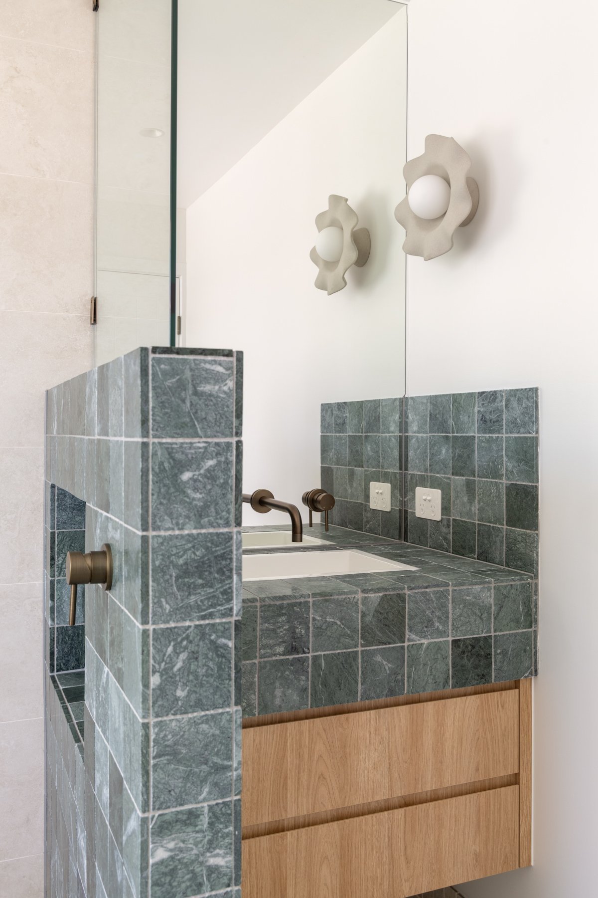 Bathroom vanity with green marble tiles, white sink, bronze faucet, large mirror, white walls, and wall-mounted gear-shaped light fixtures.