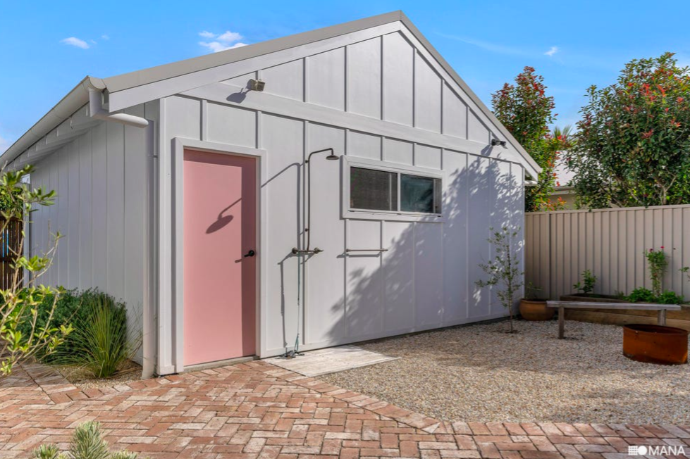 White shed with a pink door and a small window, surrounded by a brick and gravel yard, with some plants and trees