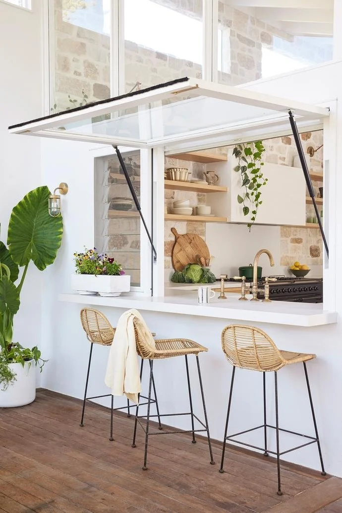 Bright kitchen with open window, wooden shelves, green plants, and bar stools with wicker seats.