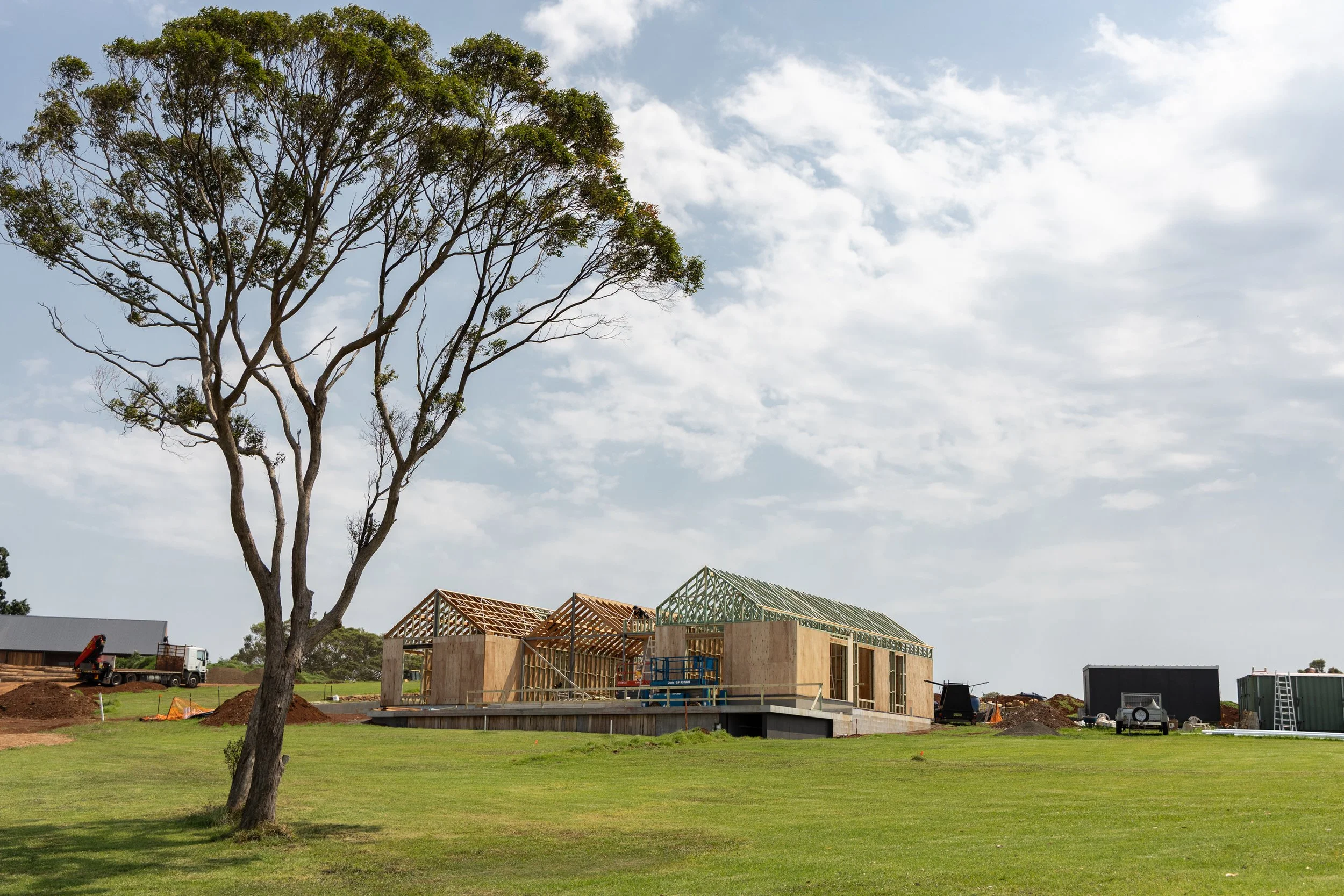 A construction site with wooden framing for a building, a large tree in the foreground, and a partly cloudy sky.