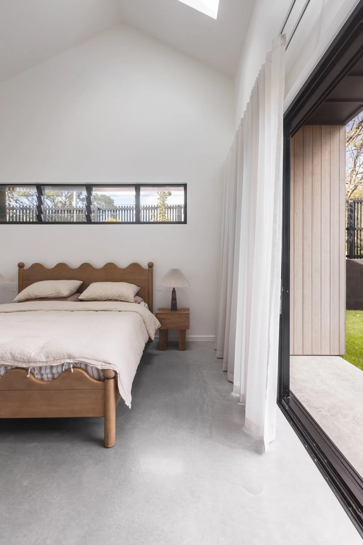 A bedroom with a wooden bed frame, beige bedding, a nightstand with a lamp, large white curtains, and an open sliding door leading outside to a patio and garden.