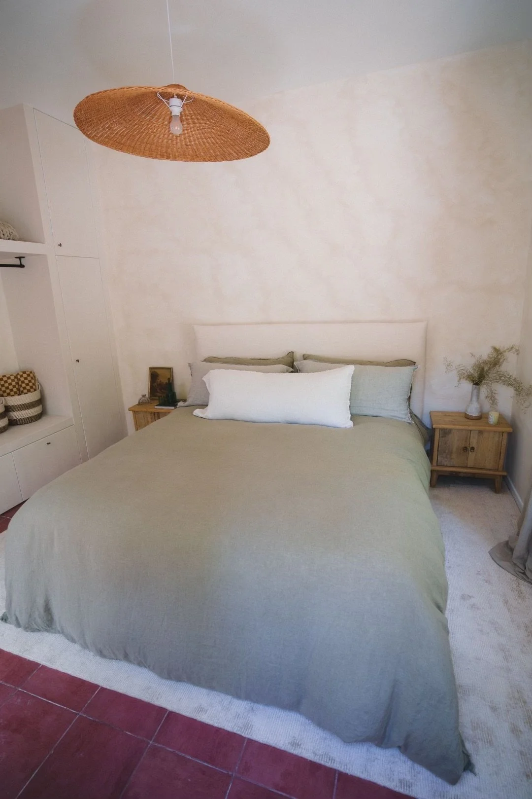 A neatly made bed with a light green bedspread, three pillows, and a beige headboard in a cozy bedroom. Two wooden nightstands flank the bed, one with a small decorative picture and the other with a vase of dried flowers. A rattan pendant light hangs from the ceiling. White cabinetry with open shelves is to the left, and a window with a curtain is partially visible on the right.