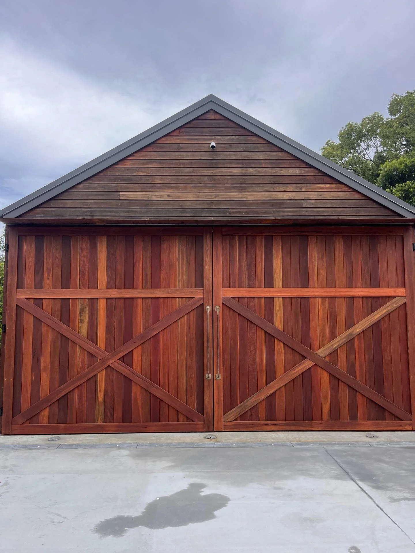 A wooden garage door with crossed patterns on it, set against a brown wooden building, with a small security camera near the top and a gray roof. The ground in front is concrete with some water stains.