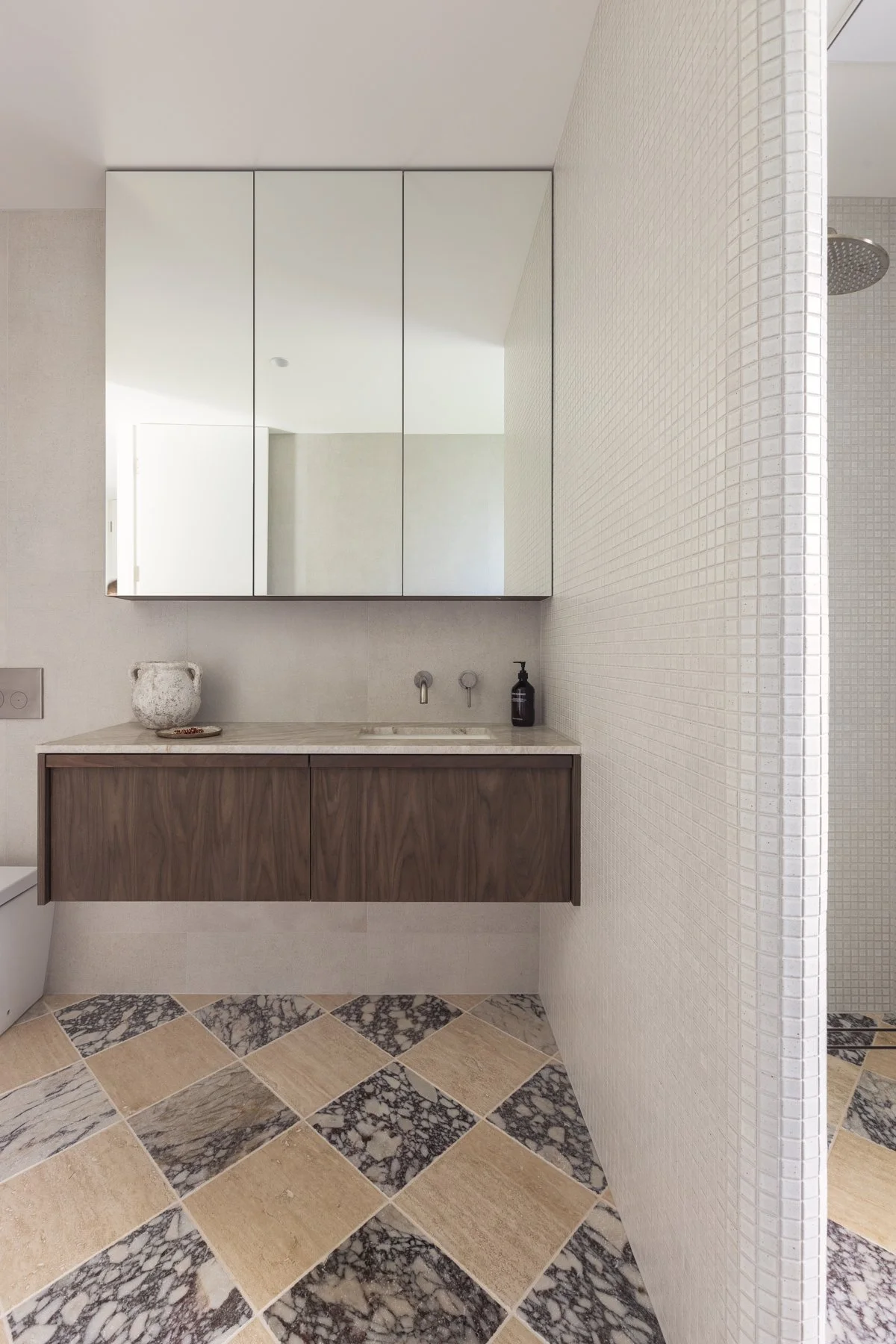 Modern bathroom with a large mirror, beige tiled floor with black and beige tiles, a floating wooden vanity, white countertop, small sink, black soap dispenser, white textured walls, and a shower area with a rain shower head.