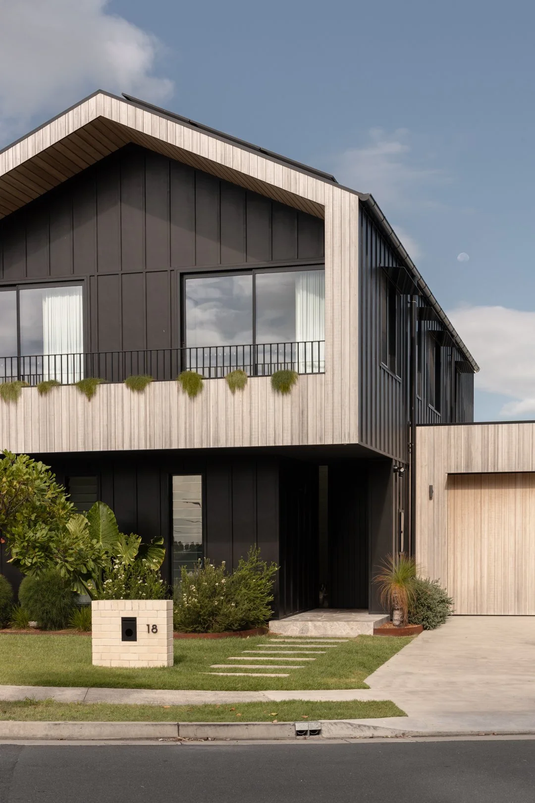 Modern two-story house with black and wooden exterior, large window, small balcony with greenery, surrounded by manicured lawn, plants, and a paved driveway, under a partly cloudy sky.