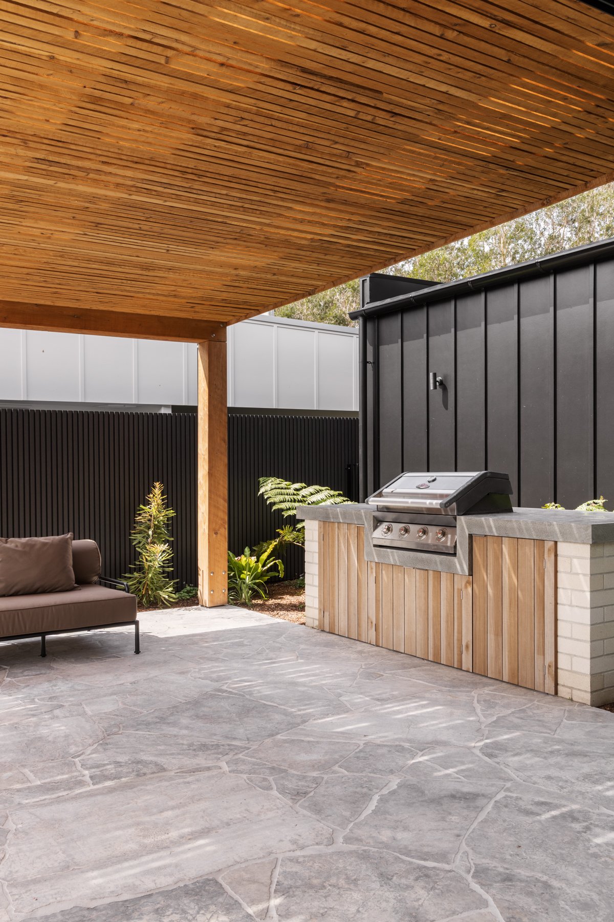 Outdoor patio with a modern built-in grill, wooden canopy, black fence, potted plants, and a seating area on a gray stone floor.