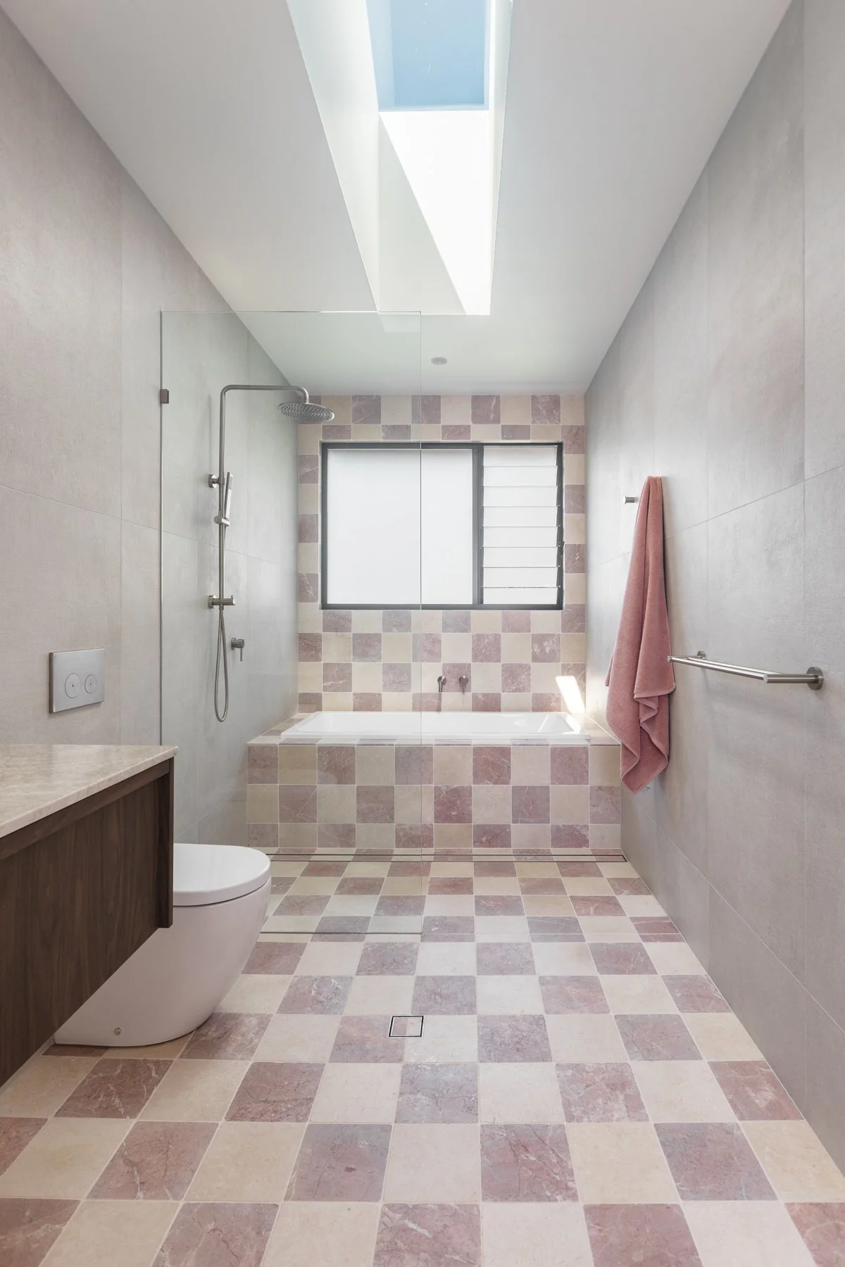 Modern bathroom with beige and pink checkered tiles on the floor and near the bathtub, a showerhead on the left wall, a window above the bathtub with frosted glass, a pink towel hanging on the right wall, a toilet in the foreground, and minimalist design with neutral tones and clean lines.