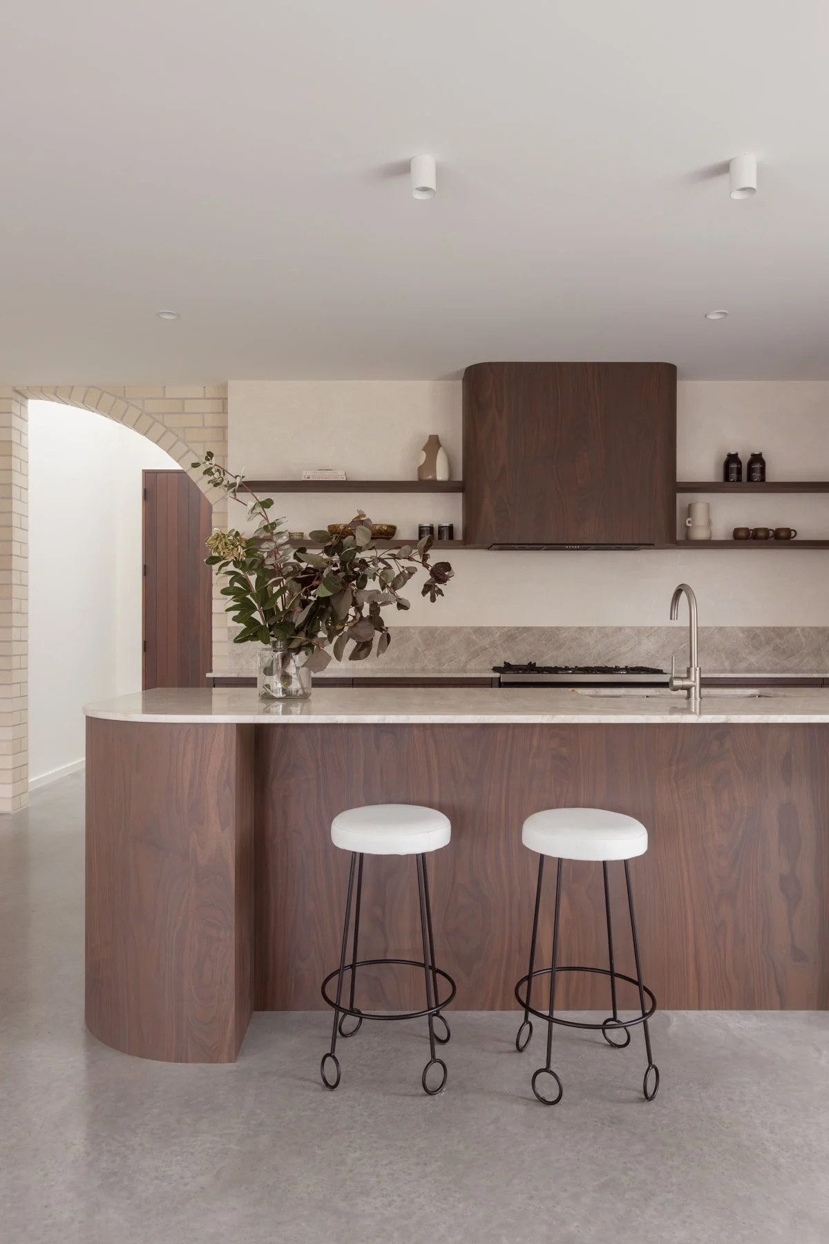 Modern kitchen with a wooden island, white marble top, and two white bar stools with black wire frames. A potted plant is on the island. Shelves with decor and a range hood are in the background, along with an archway and brick wall.