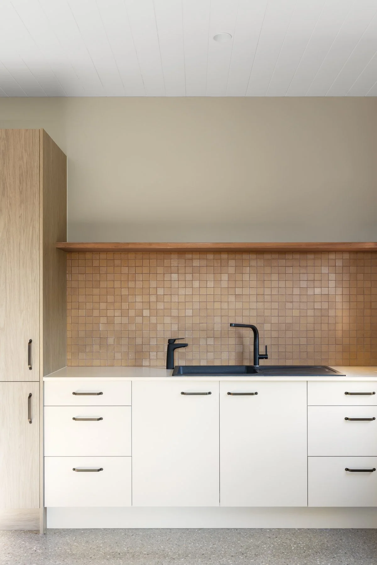 Modern kitchen with white cabinets, black sink and faucet, beige tiled backsplash, and wooden upper shelf against a beige wall, with a white ceiling.