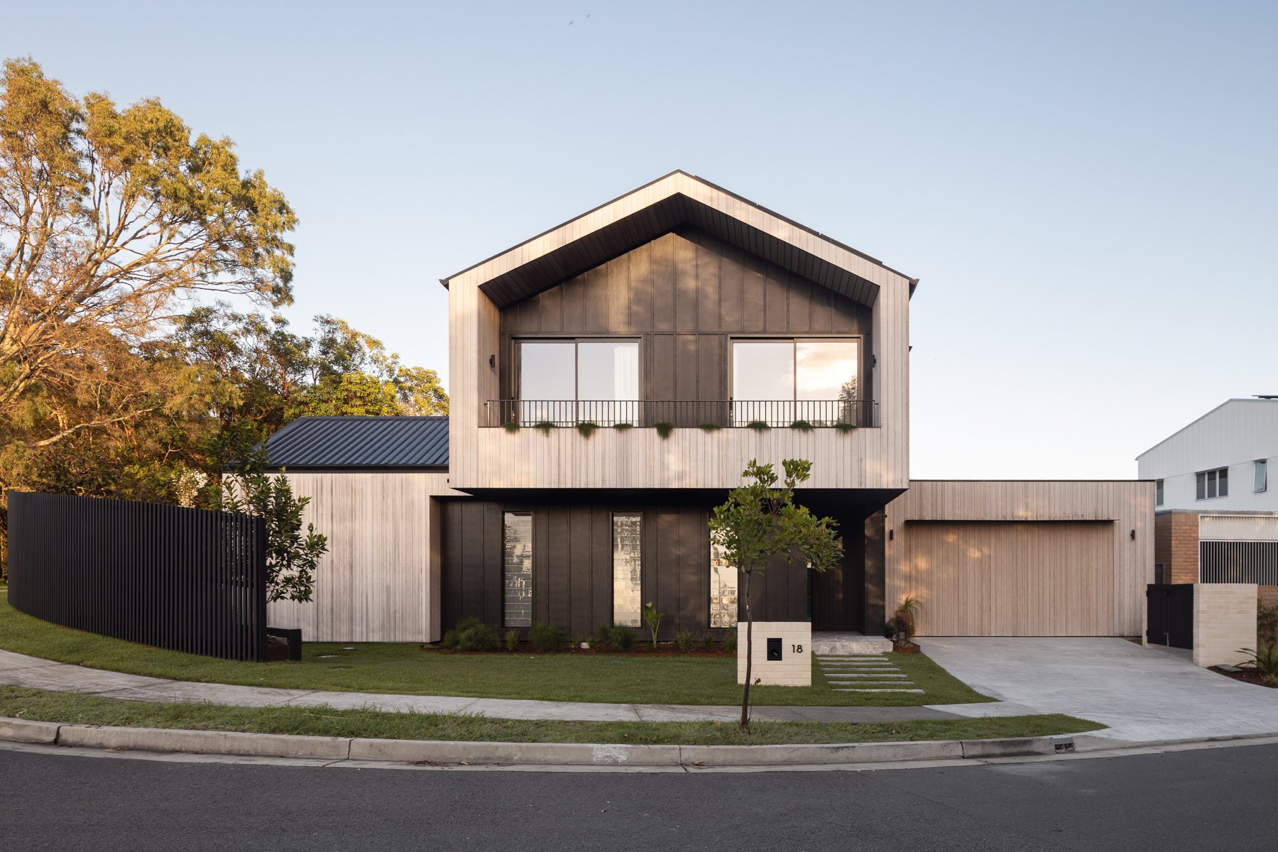 Modern two-story house with wood and black exterior finishes, large windows, small front yard with a tree, and a driveway, on a suburban street.