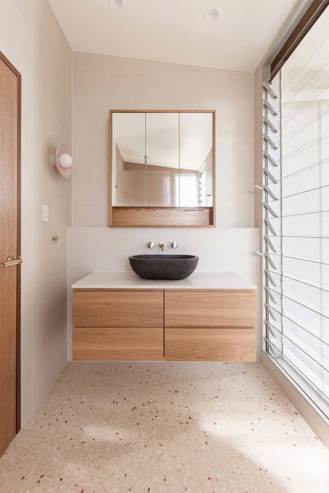 Minimalist bathroom with a floating wooden vanity, a black stone vessel sink, a wall-mounted faucet, a mirror with a wooden frame, and a large window with blinds.