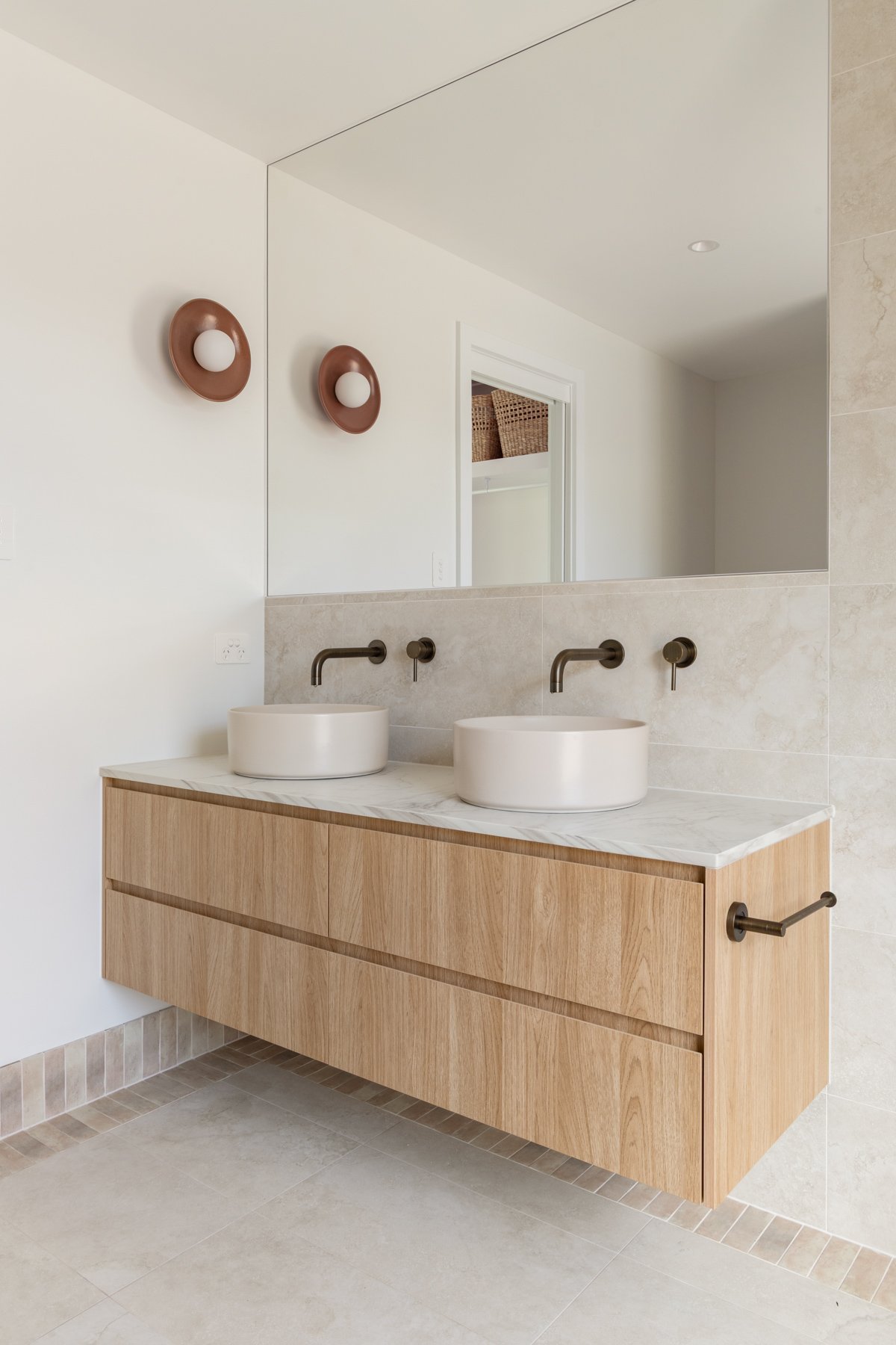 Modern bathroom with a double vanity featuring two round vessel sinks, wall-mounted bronze faucets, a large mirror, and a wooden cabinet. Two decorative wall lights are visible on the mirror, and the room has beige tiled walls and floor.