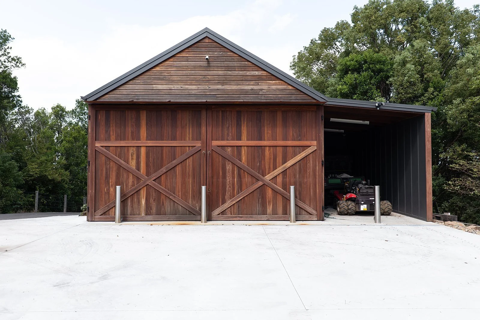 A wooden garage with two sliding doors on the left and a partially open side showing a riding lawn mower inside. The garage is on a concrete driveway with a green background of trees.