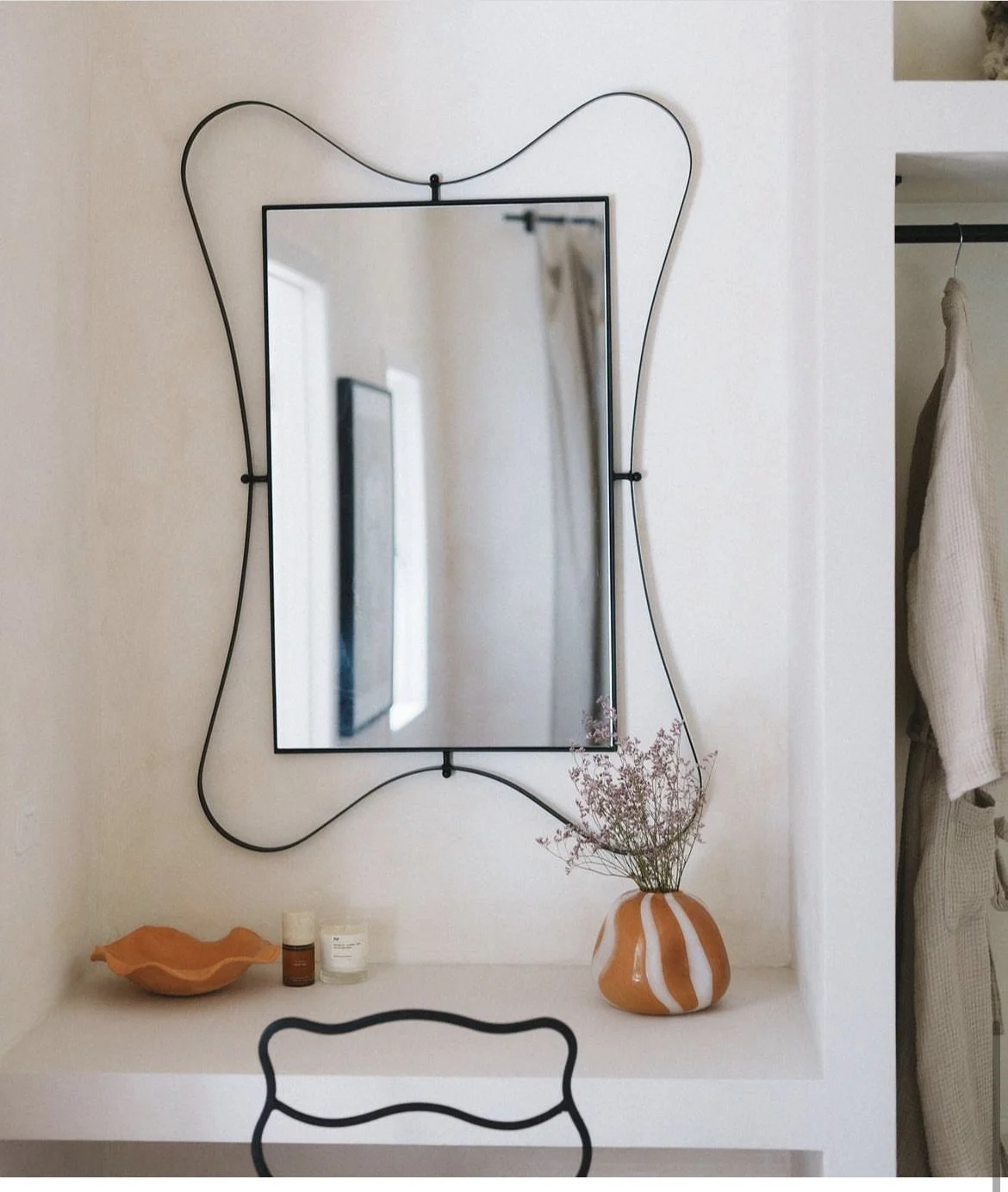 Decorative wall mirror with black metal frame and wavy border, mounted on a white wall. Below the mirror, a white surface holds an orange ceramic bowl, a small amber glass bottle, white candles, and a white and orange striped vase with dried pink flowers. To the right, an open closet with a white garment hanging.