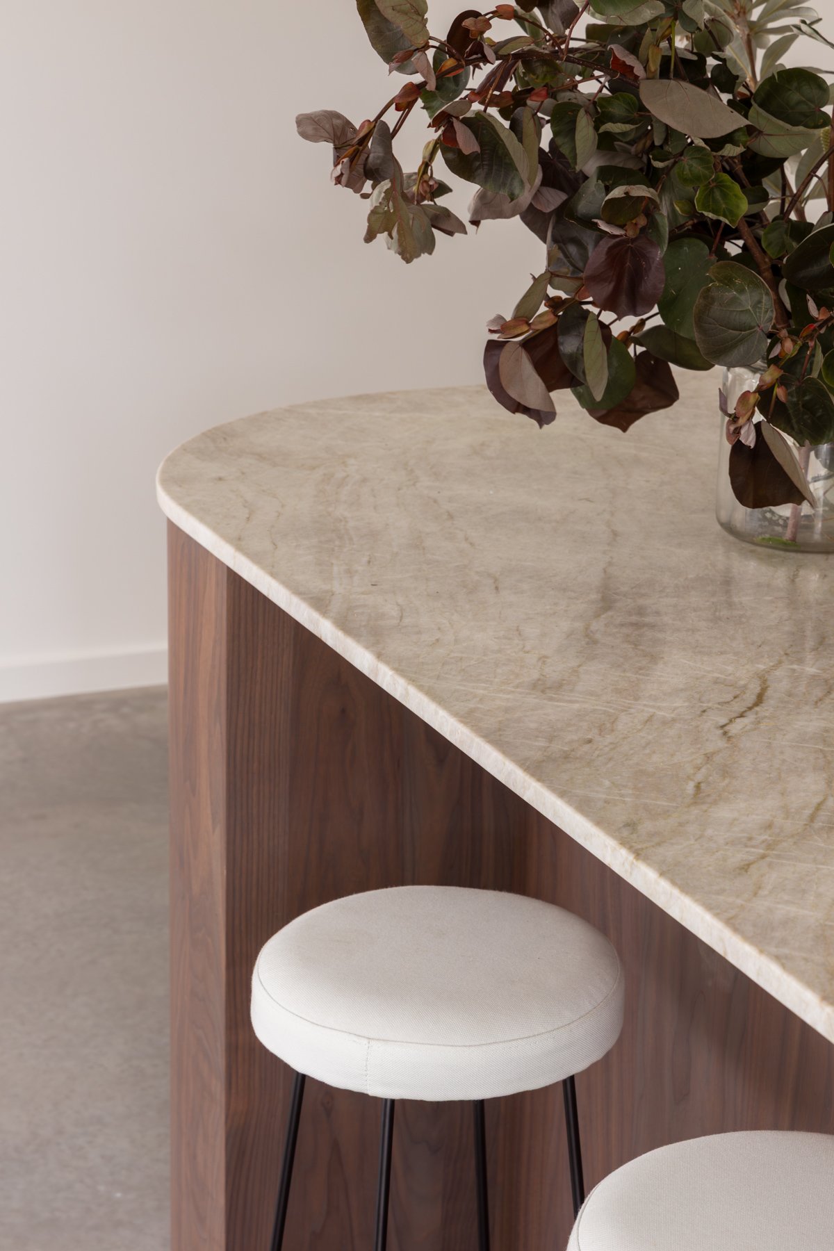 A beige marble top bar or counter with a wooden front panel, paired with two white cushioned bar stools with black metal legs, and a glass vase containing a leafy green and purple plant.