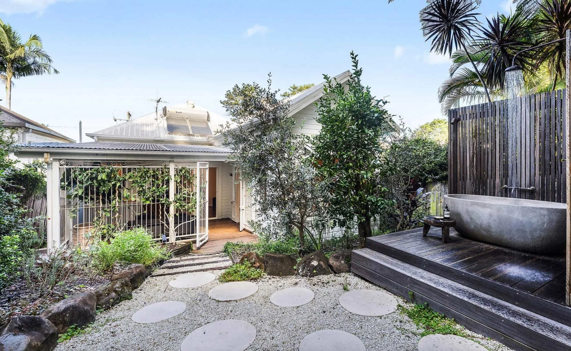 A backyard garden area with a stepping stone pathway, lush green plants, and trees, enclosed by a white house with a metal roof and a wooden fence with a outdoor shower. There is a large concrete bathtub on a wooden deck with a bottle and a small tray on it.