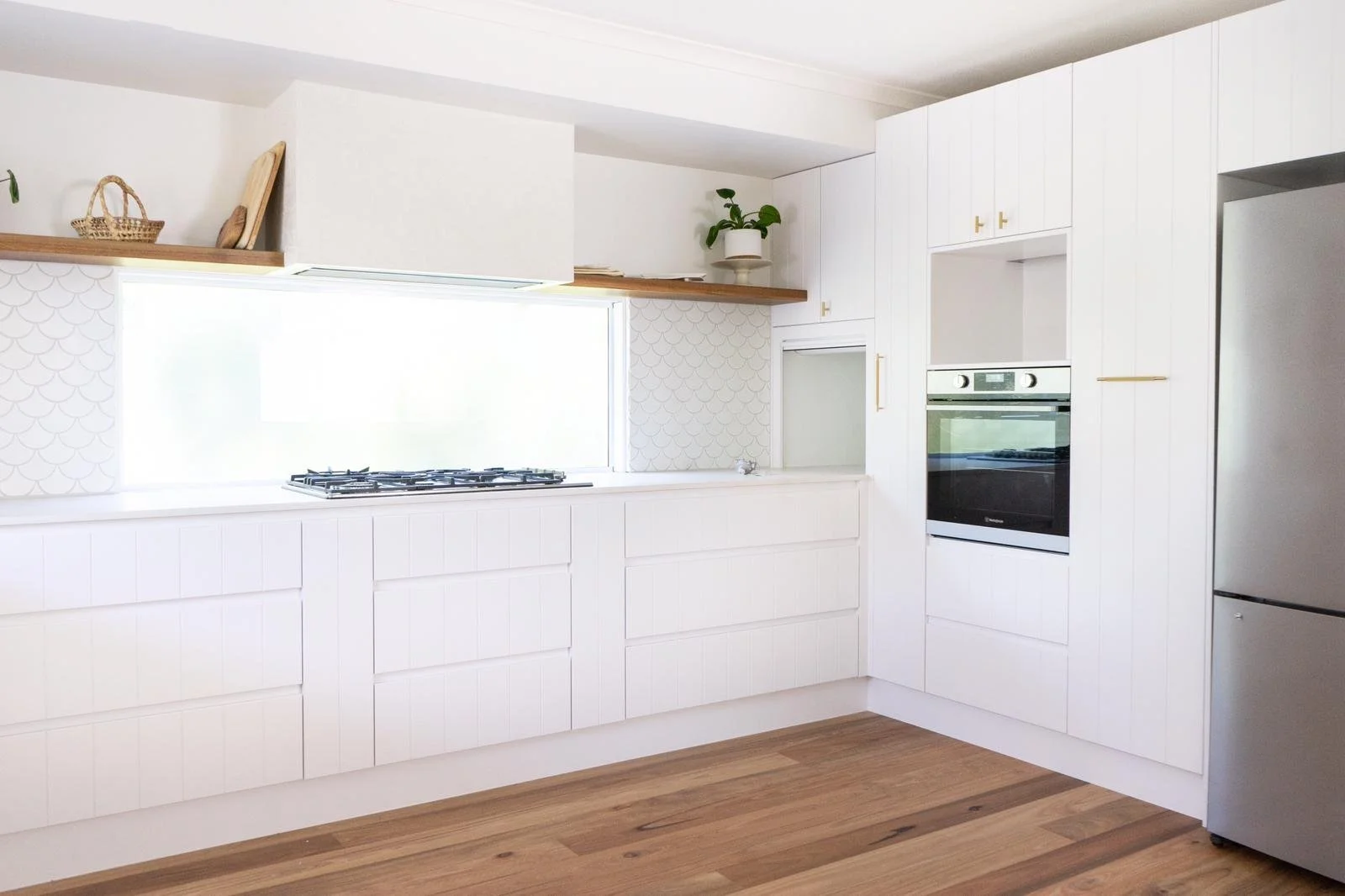 Modern white kitchen with built-in oven, stove, refrigerator, white cabinets, wood accents, and hardwood flooring.