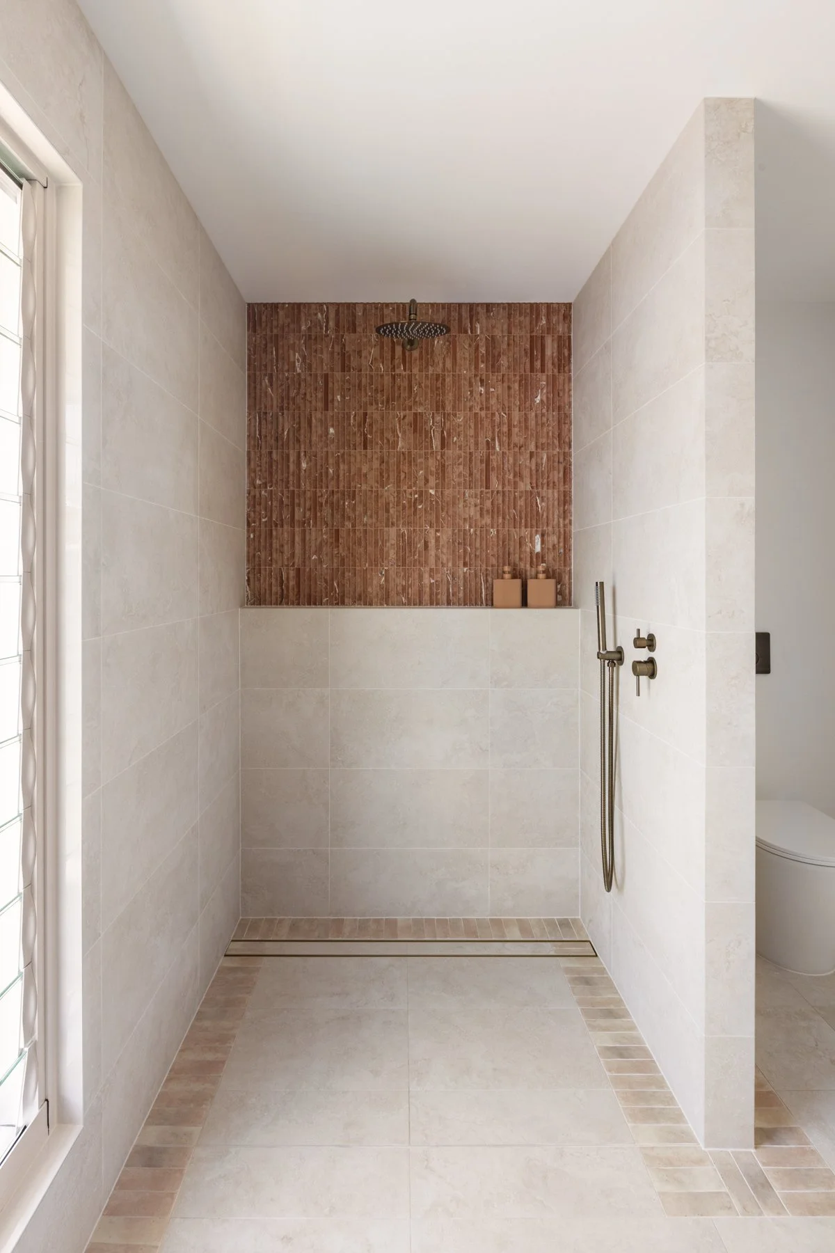 Modern walk-in shower with beige tiles, a brick-red accent wall, a rain showerhead, and a handheld shower fixture.