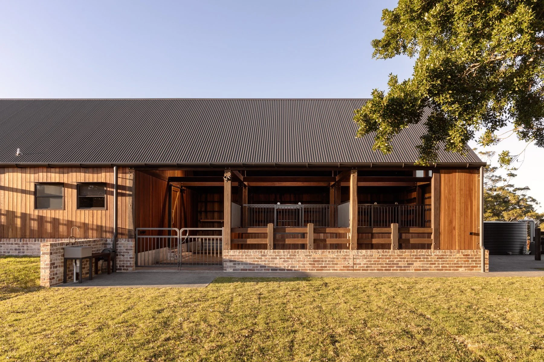 A barn or similar structure with a metal roof, wooden walls, and a brick foundation. The building has open sections with railings and stairs leading up to a deck area. There's greenery and a tree on the right side, and a clear blue sky above.