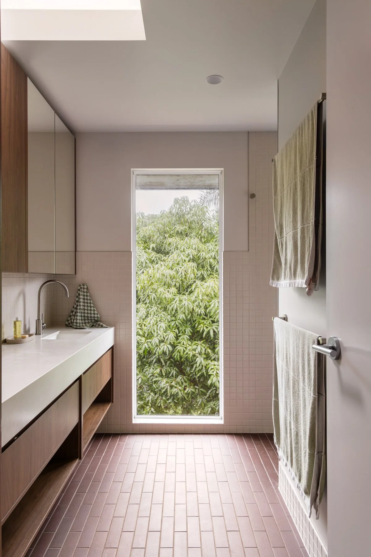 Modern bathroom with large window showing green foliage outside, beige tiled floors, white wall tiles, wooden vanity with sink, towels hanging on a wall-mounted rack, and natural light.