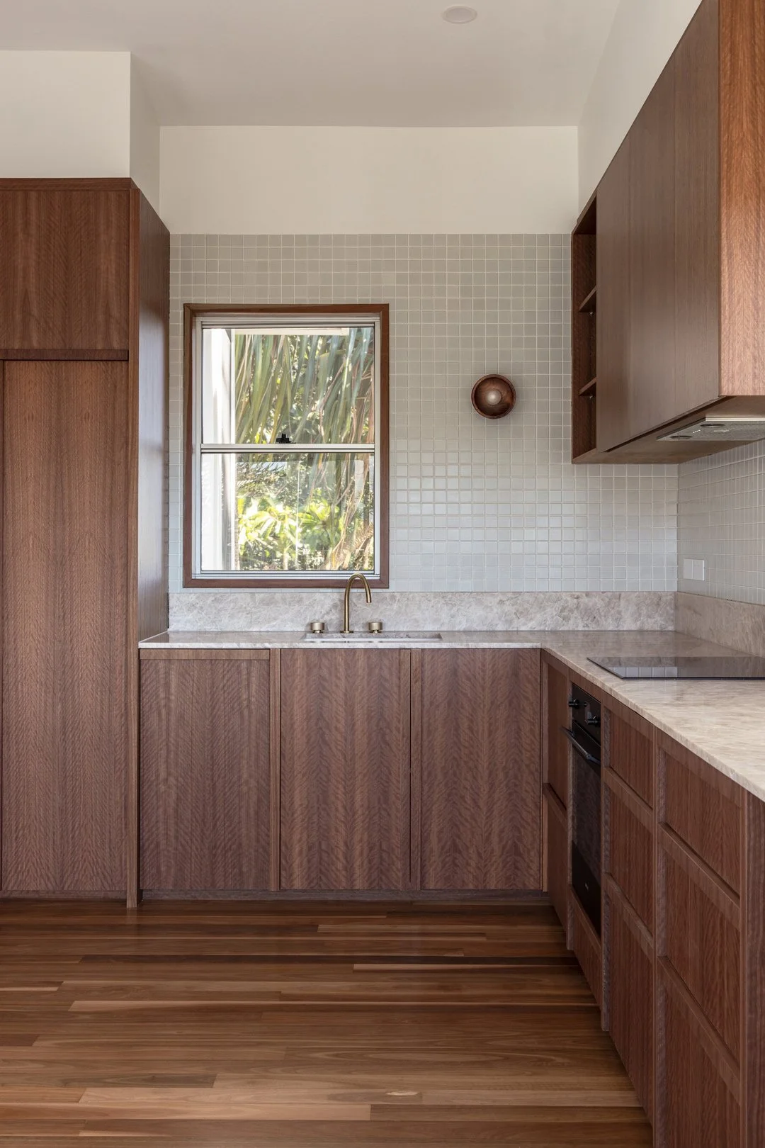 A kitchen with wooden cabinets, a window above the sink, a marble countertop, and a built-in stove. The walls have small white tiles and there is a world globe on the wall to the right of the window.