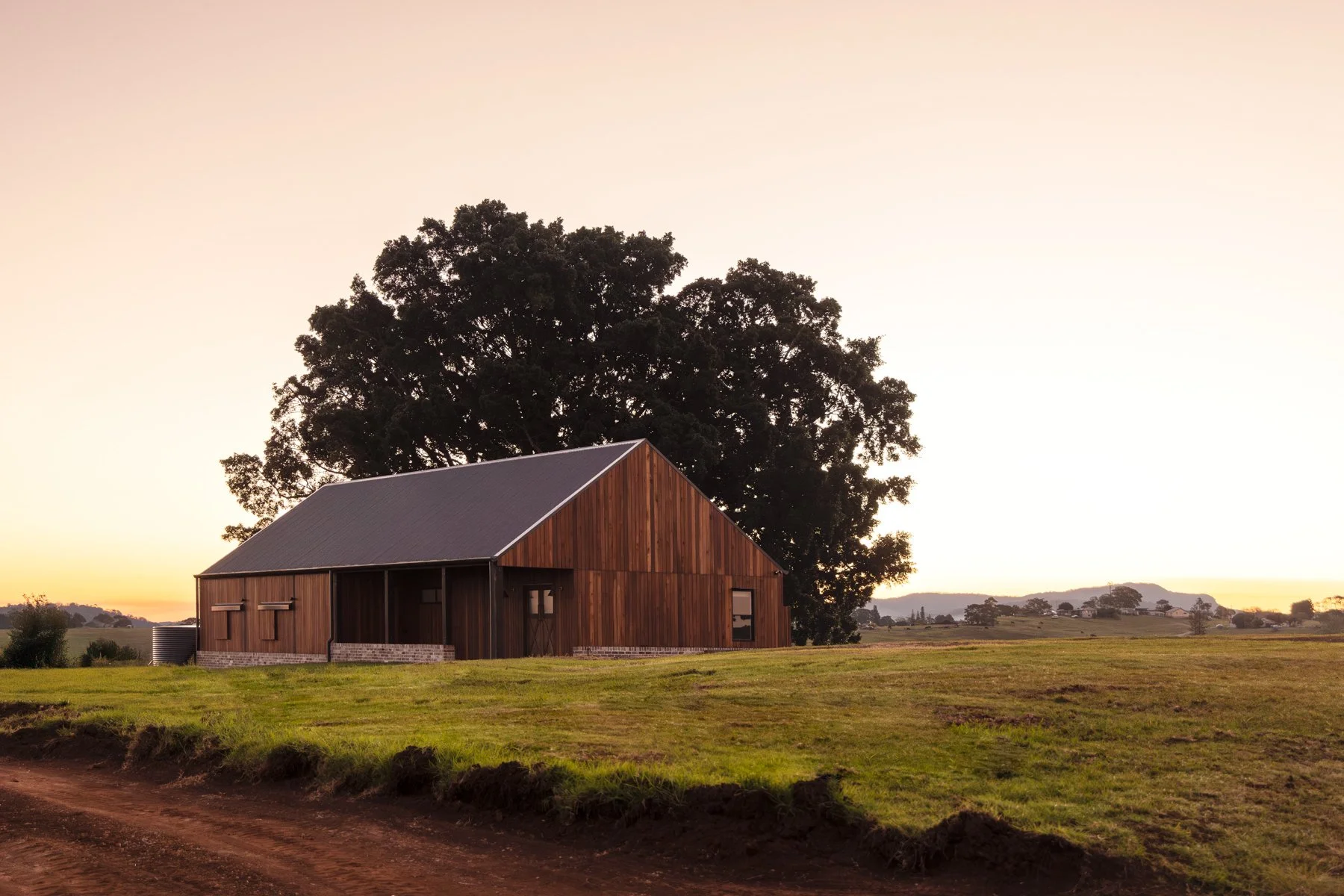 A wooden barn on a grassy field with a large tree behind it, during sunset with a clear sky and distant landscape.