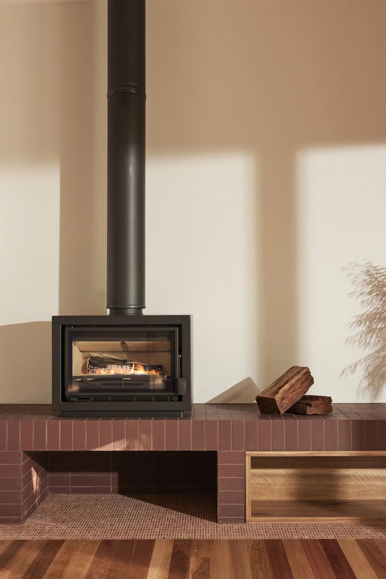 A modern wood-burning stove with a black chimney pipe in a cozy living room, placed on a brick hearth with firewood nearby and a wooden shelf underneath.