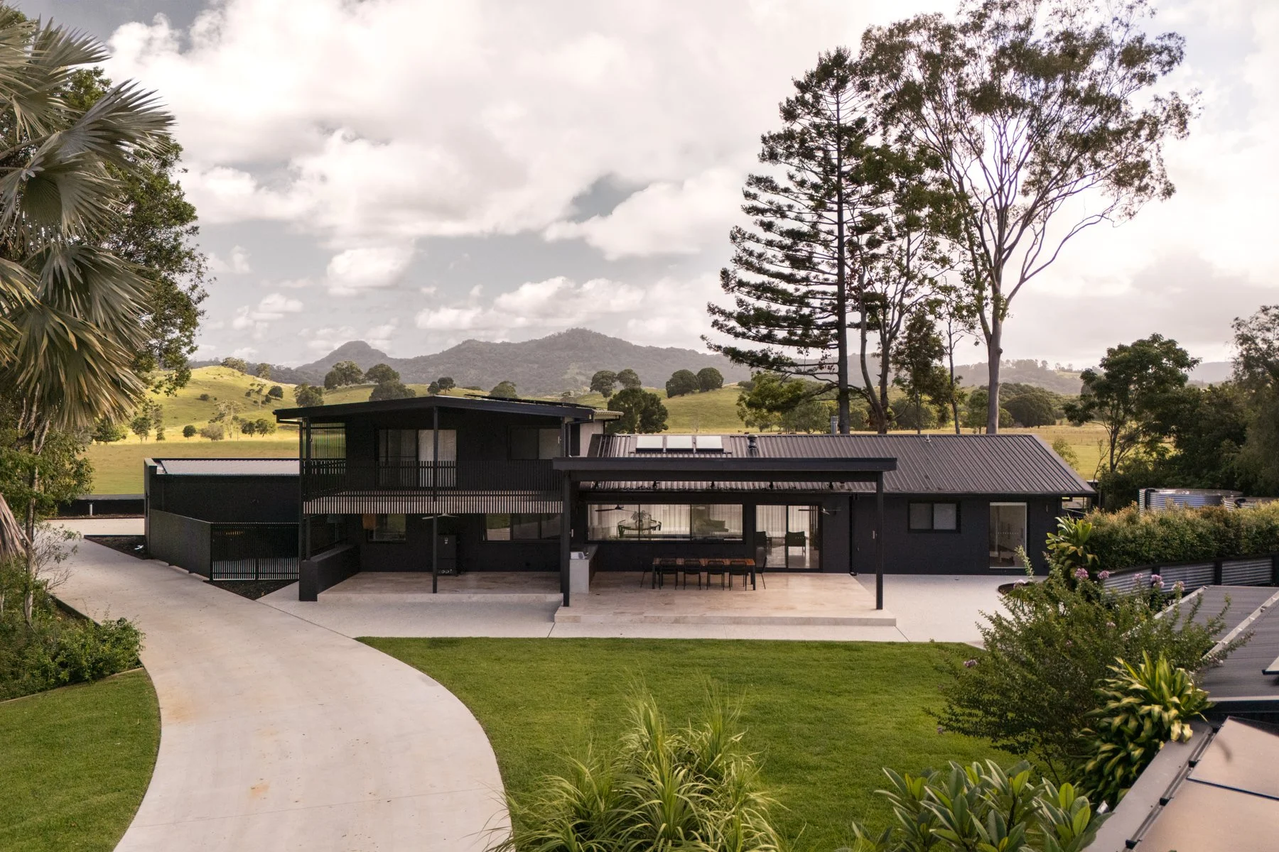 A modern black house with large glass windows and a sloped metal roof, situated in a lush green landscape with rolling hills and tall trees in the background, under a partly cloudy sky.