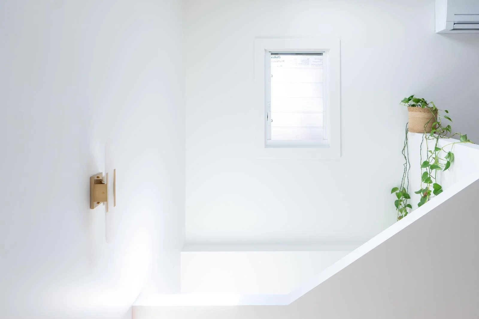 Minimalist white room with a small window, wooden wall light fixture, and green plants on a white ledge.