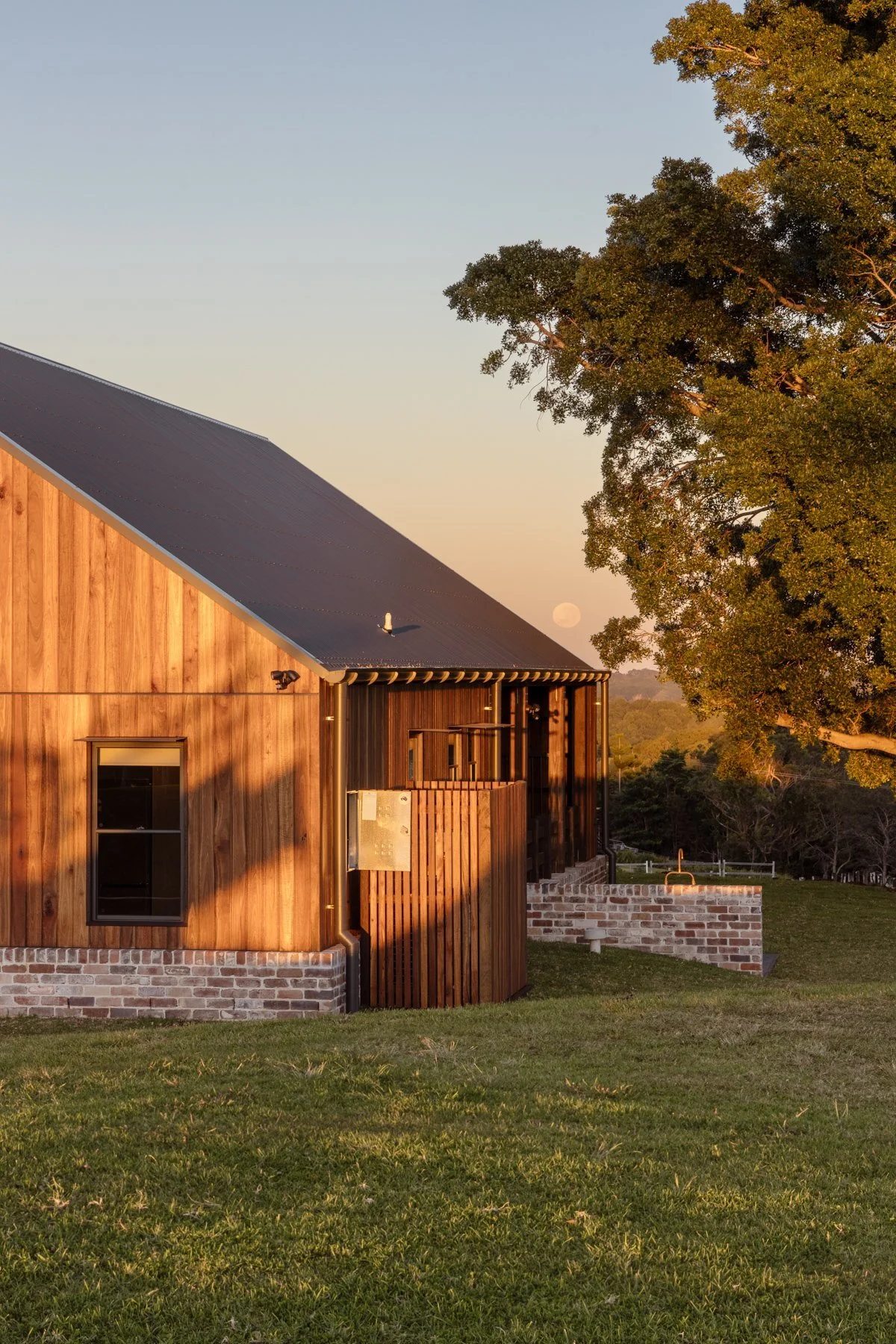 A modern wooden house with brick foundation, set in a grassy yard, with a large tree and the moon visible in the clear sky during sunset.
