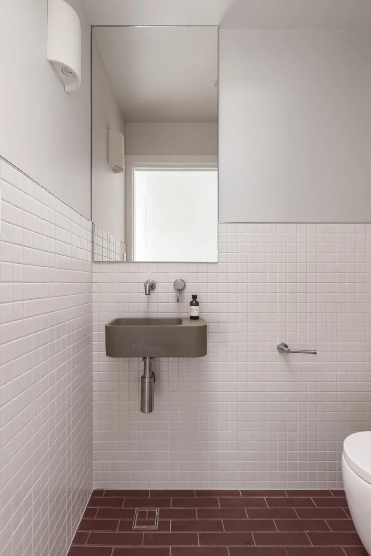 Small bathroom with white tile walls and dark red tile floor, featuring a wall-mounted sink, large mirror, and window with natural light.