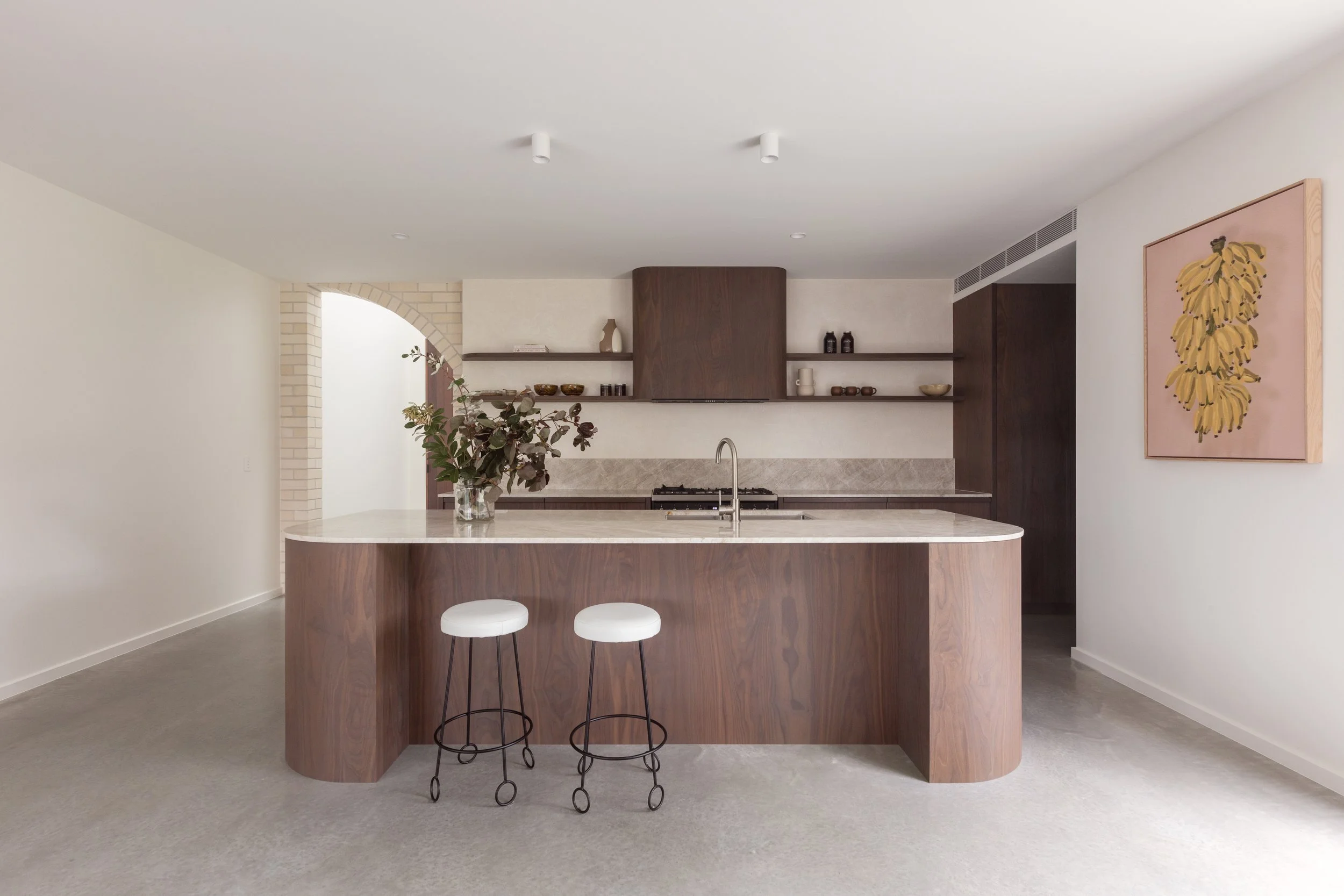 Modern kitchen with a curved wooden island, two white barstools, open shelving with dishes and decor, and a large wall art piece of yellow bananas.