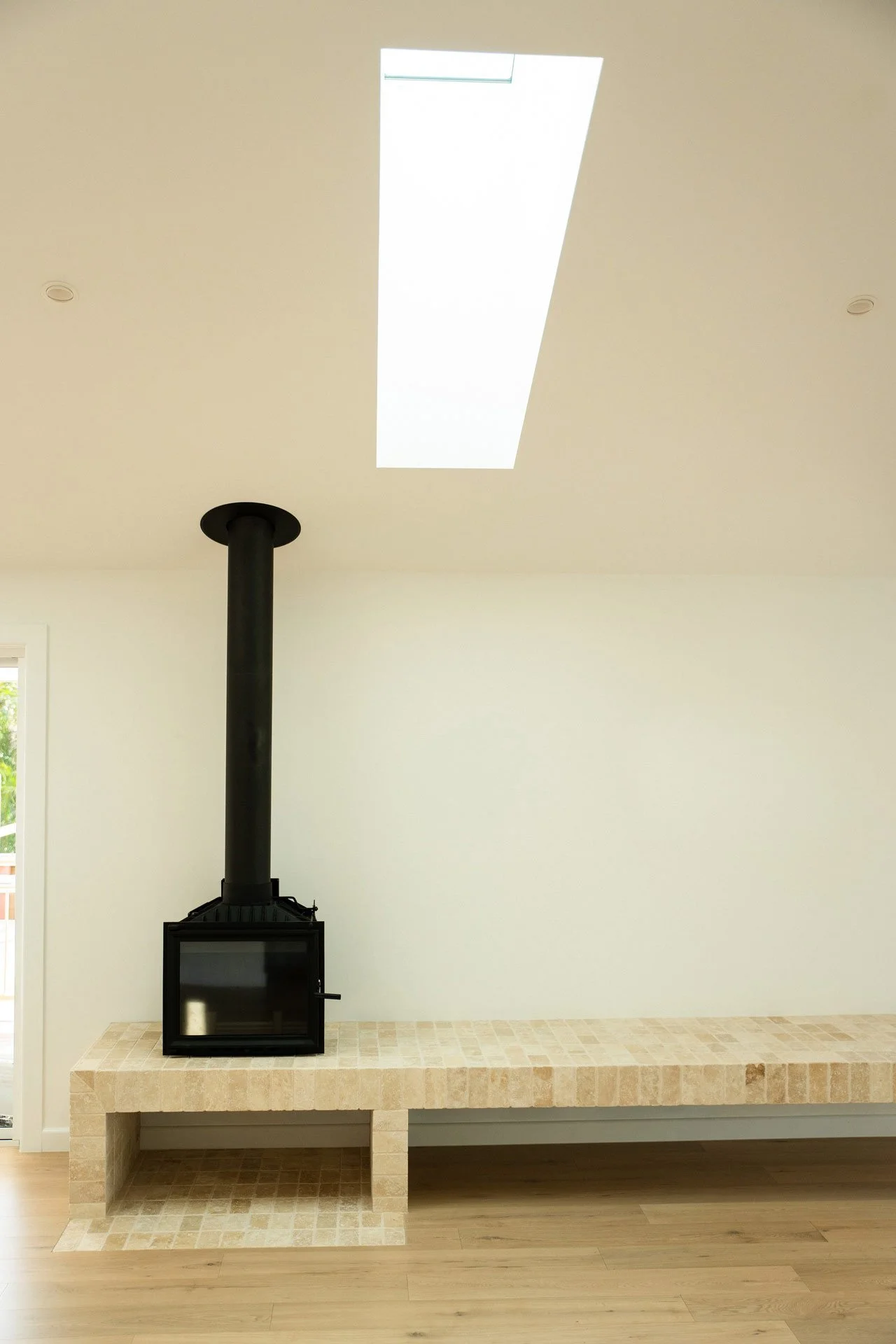 A black stove with a glass door and a chimney pipe on a tiled hearth in a room with white walls and a large, rectangular skylight ceiling.